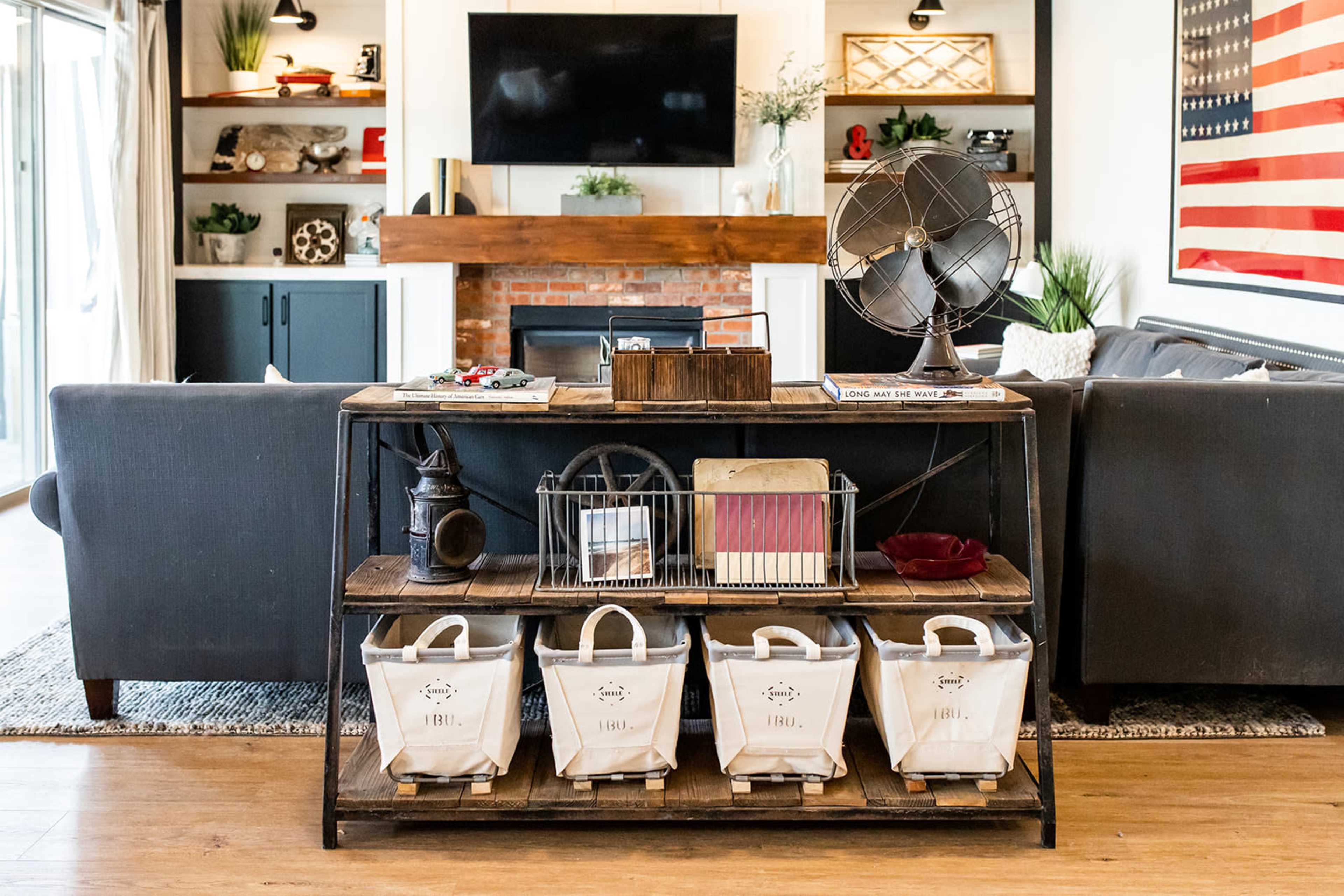 The image depicts a cozy living room with a rustic wooden console table displaying books and decorative items, accompanied by a fan and several storage bins beneath it, framed by a fireplace and a television on the wall.