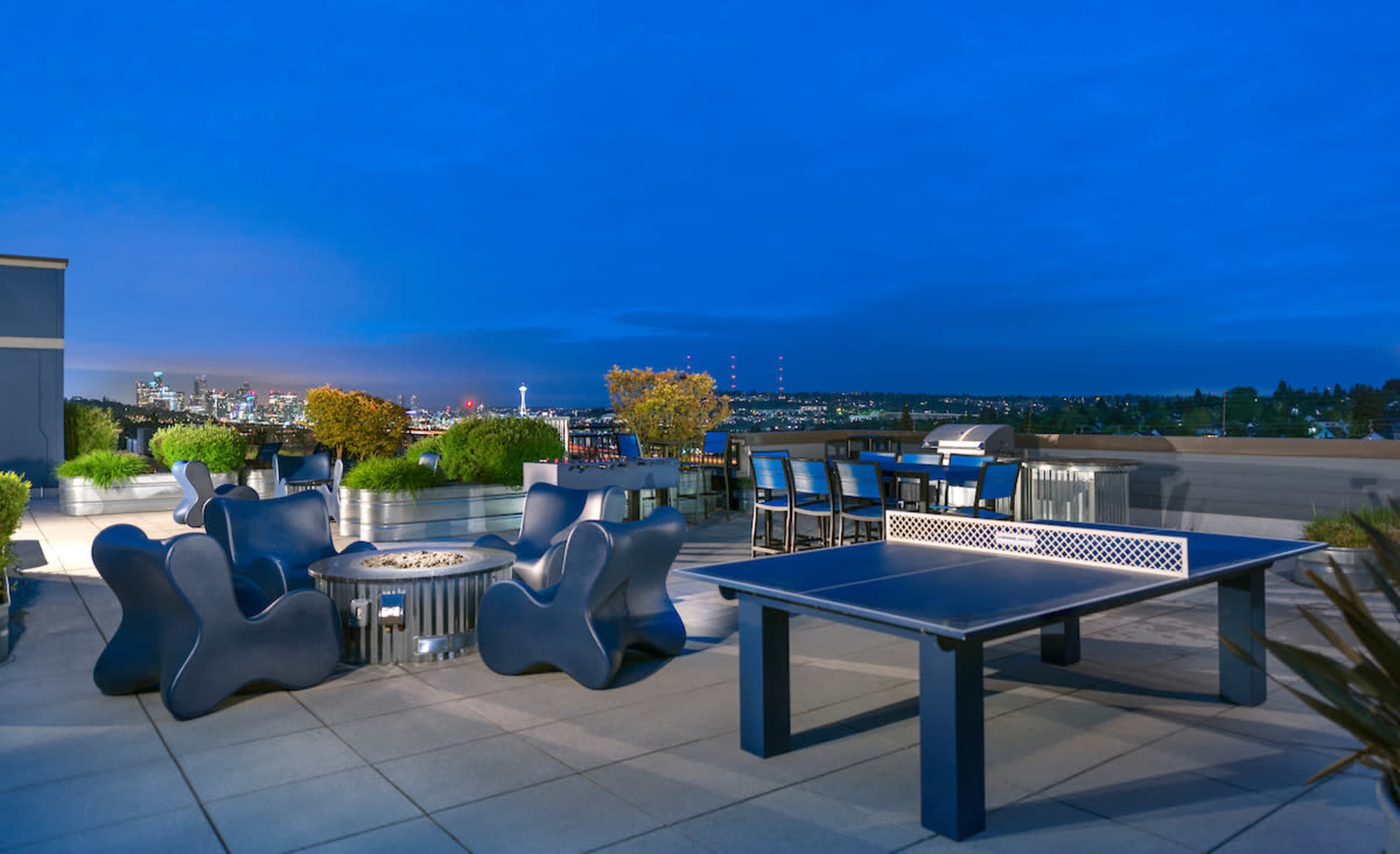 A rooftop terrace featuring a ping pong table, modern seating arrangements, and a view of a city skyline at dusk.