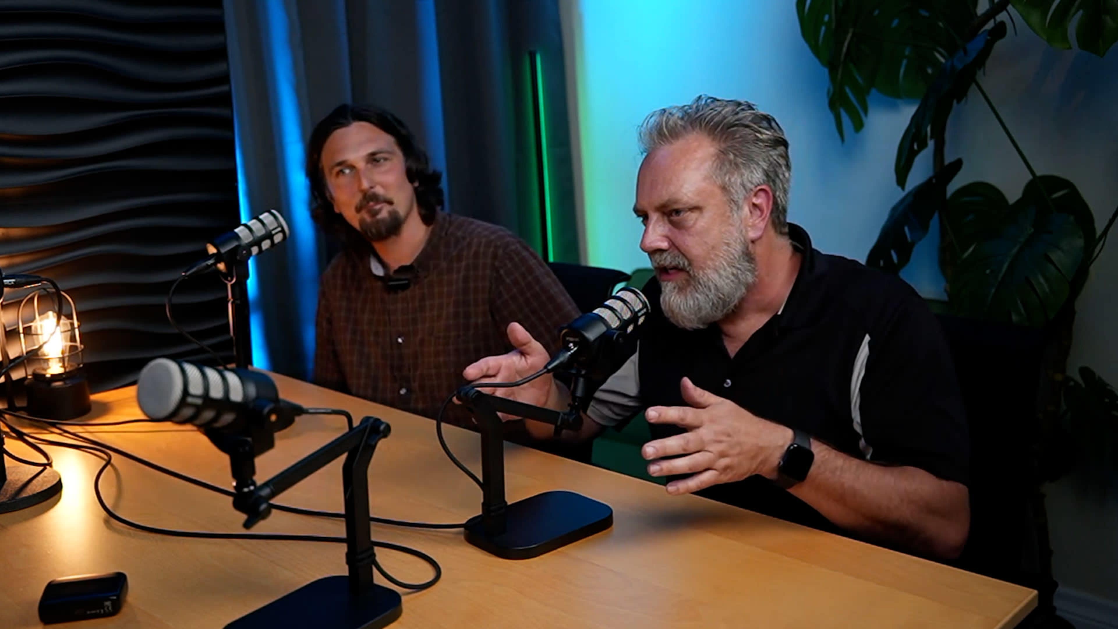 Two men are seated at a table with microphones, engaging in a conversation against a backdrop of illuminated curtains and a plant.