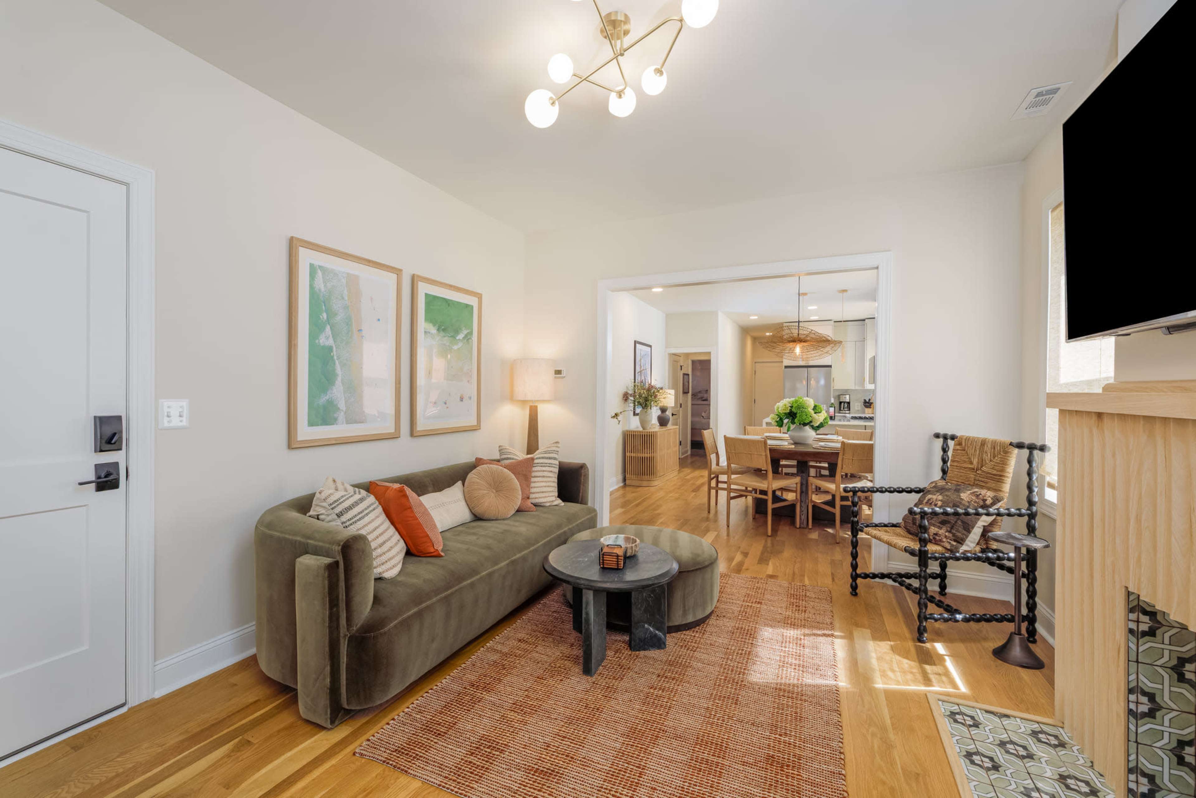 The image shows a cozy living room featuring a green couch, a round coffee table, and framed artwork on the walls, with an open doorway leading to a kitchen area.