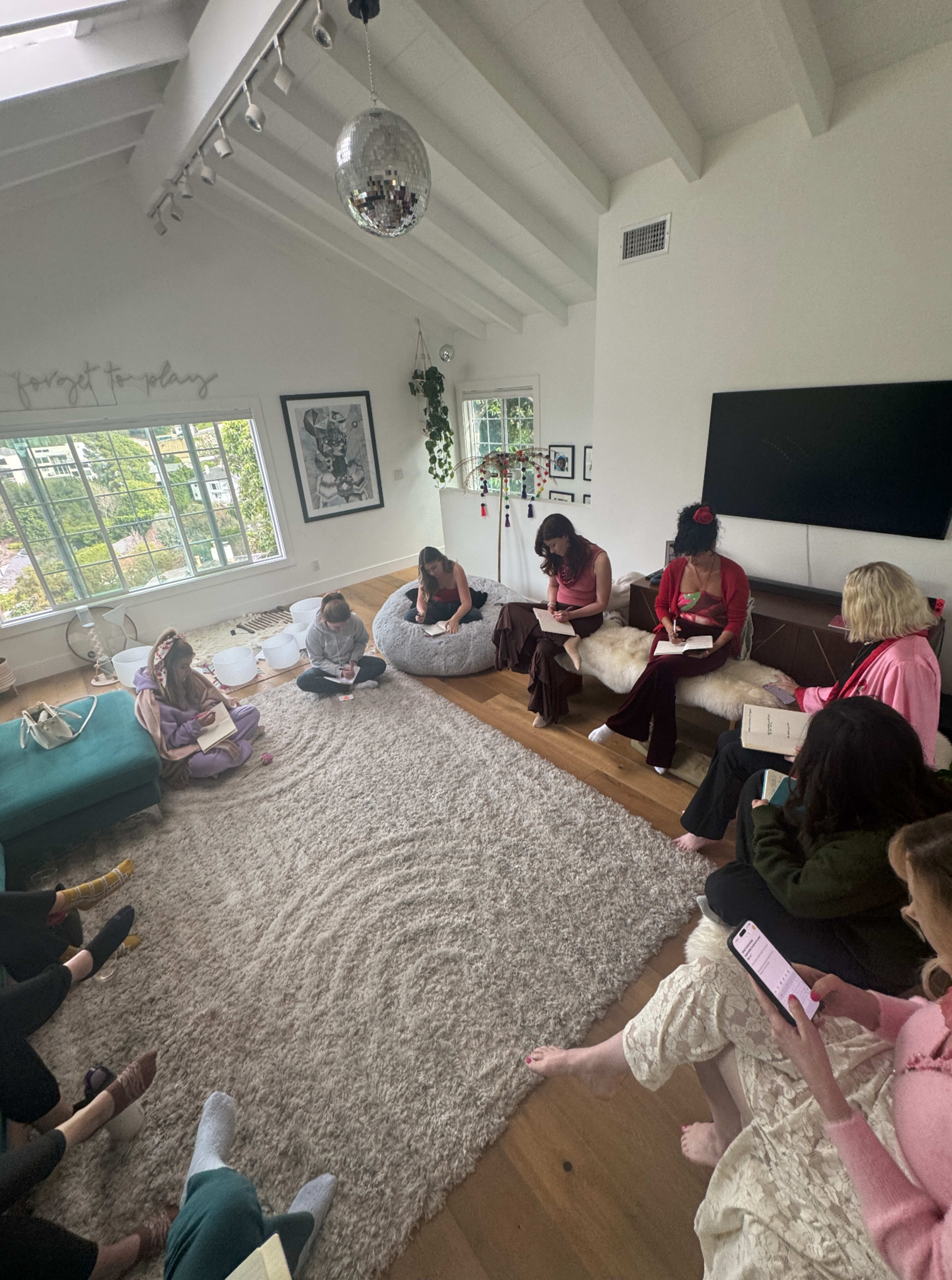 A group of people sits in a circle on a carpeted floor in a well-lit room, engaged in note-taking and discussion.