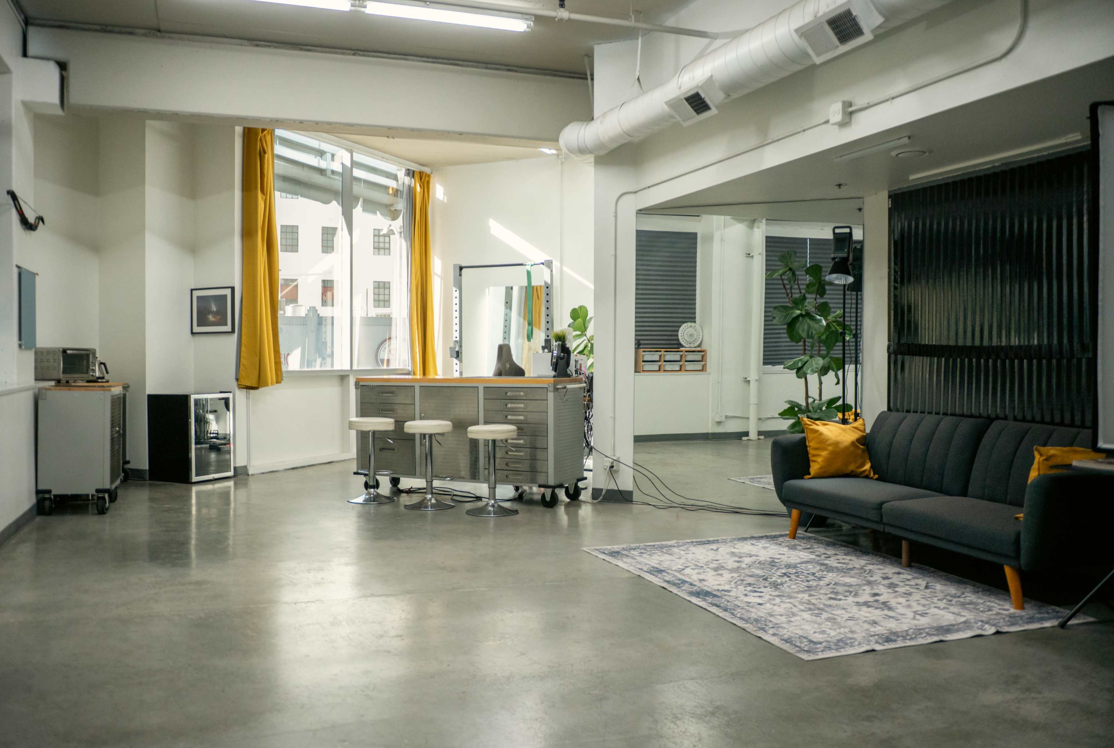 The image shows a modern and spacious office interior featuring a gray sofa, a small kitchen area, and large windows with yellow curtains.