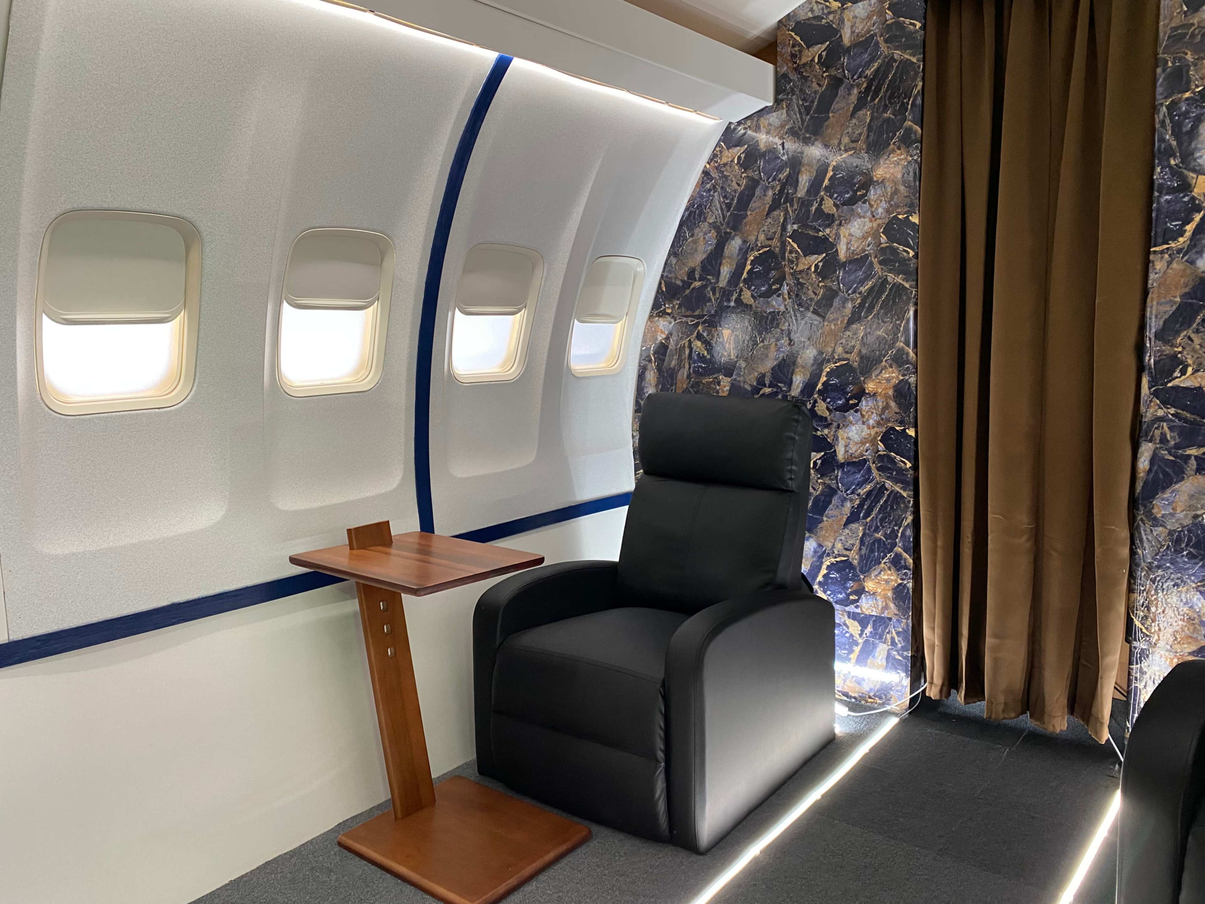A seating area inside an airplane, featuring a black chair next to a small wooden table with large windows and a decorative wall.