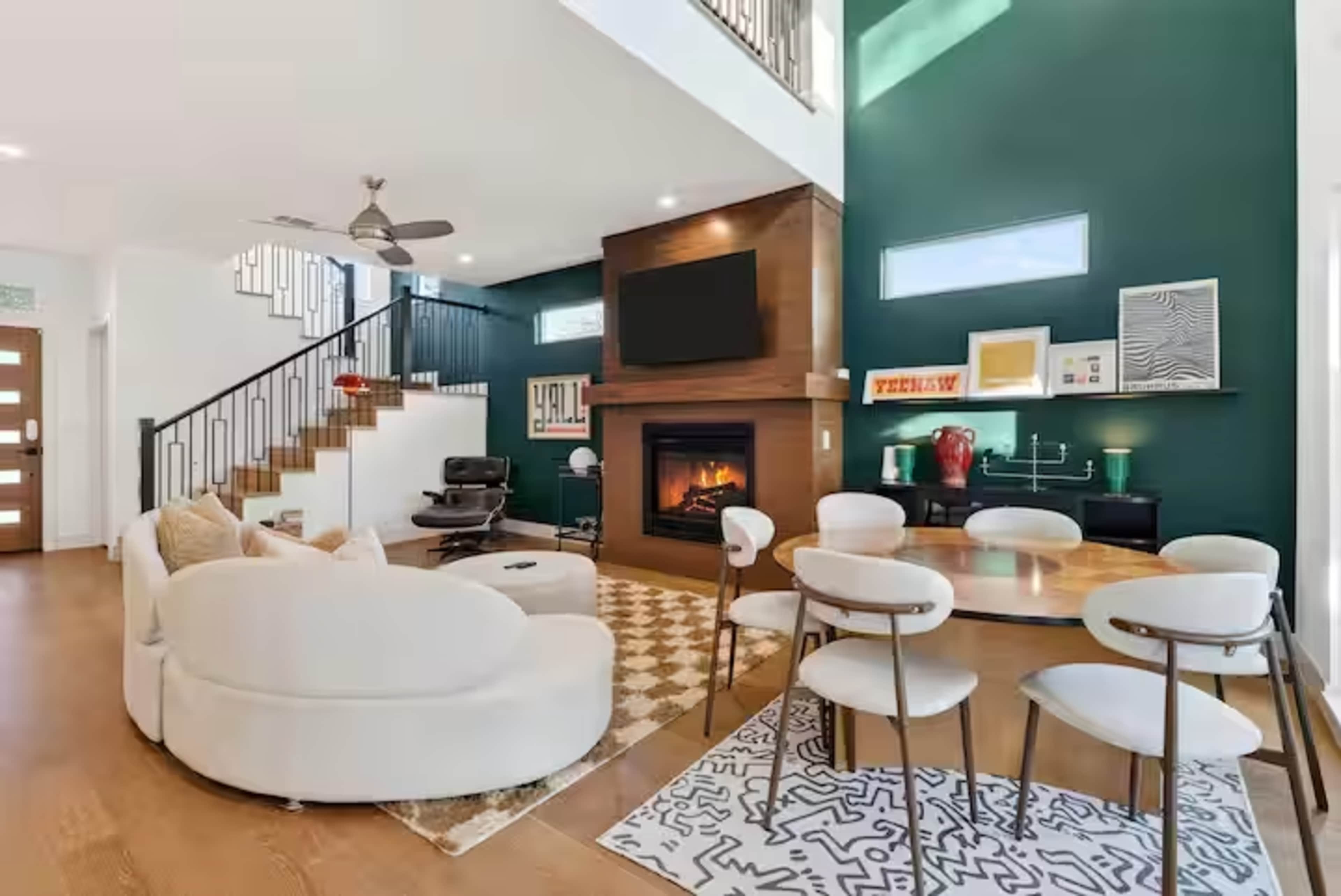 A modern living room features a green accent wall, a fireplace with a TV mounted above it, and a combination of plush seating and a dining area.