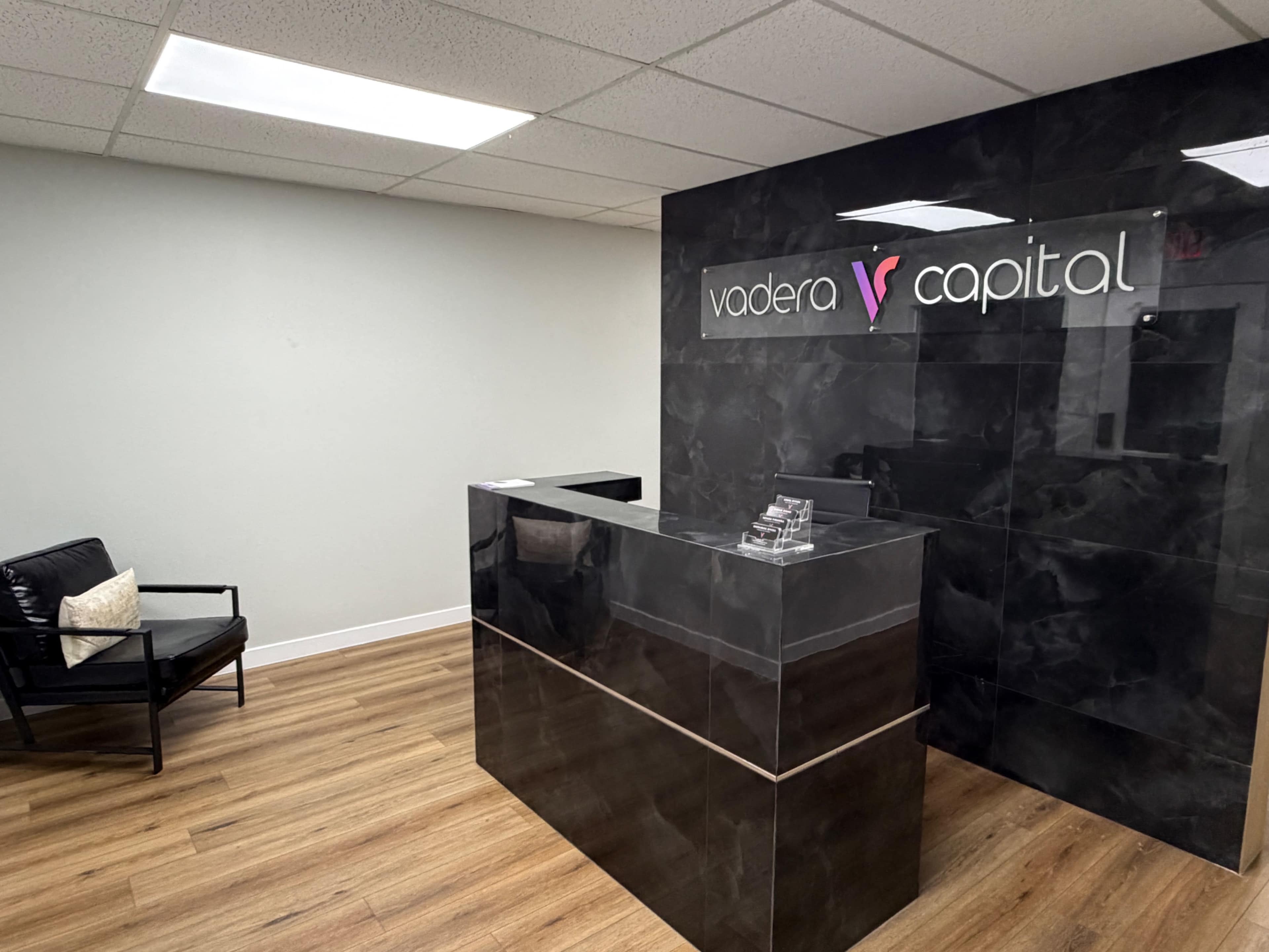 The image shows a modern reception area featuring a black marble counter with a sign that reads "vadera capital" and a black chair against a light-colored wall.