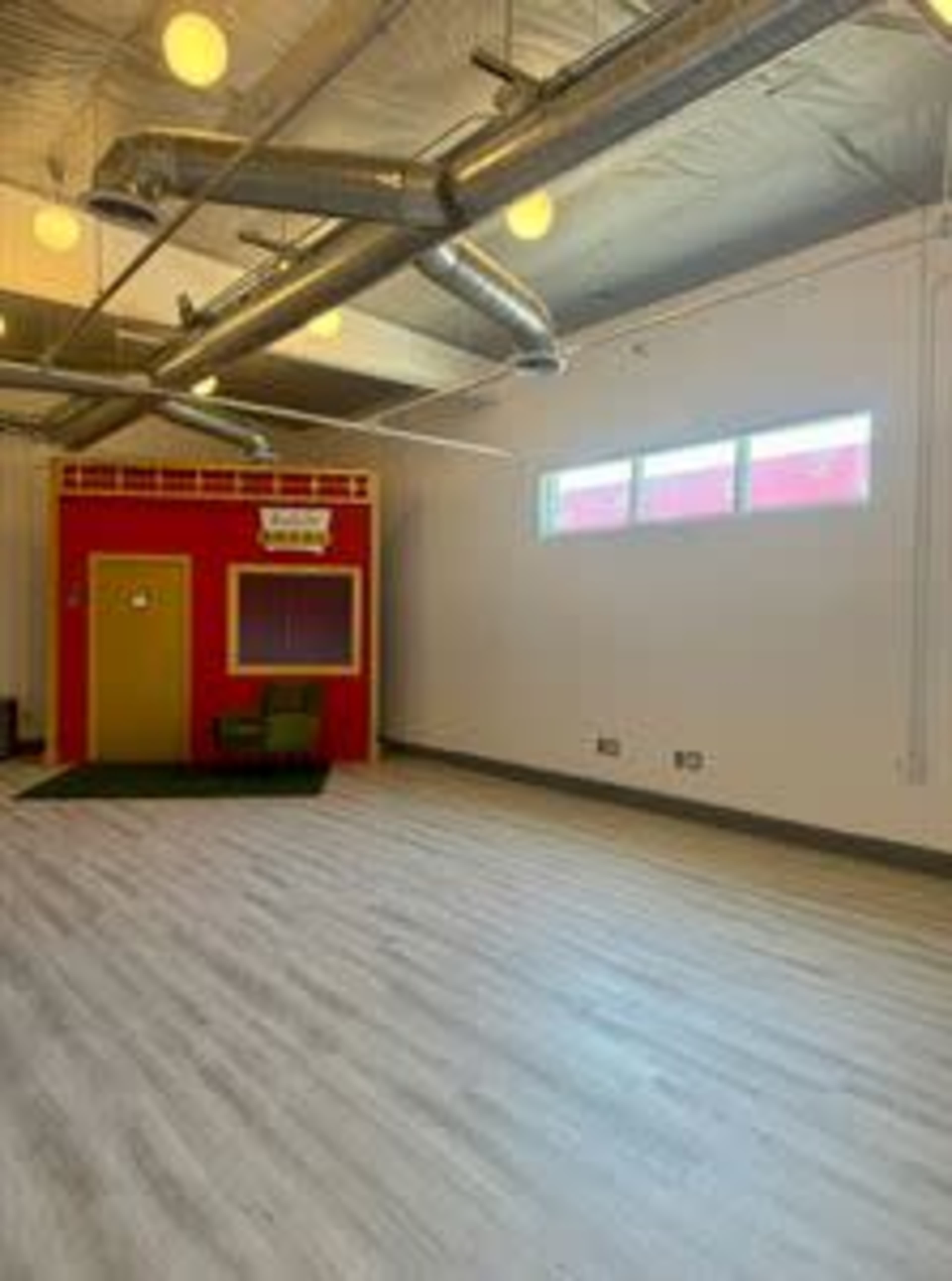 A spacious room with wooden flooring, featuring a red structure resembling a small building, and two windows letting in natural light.