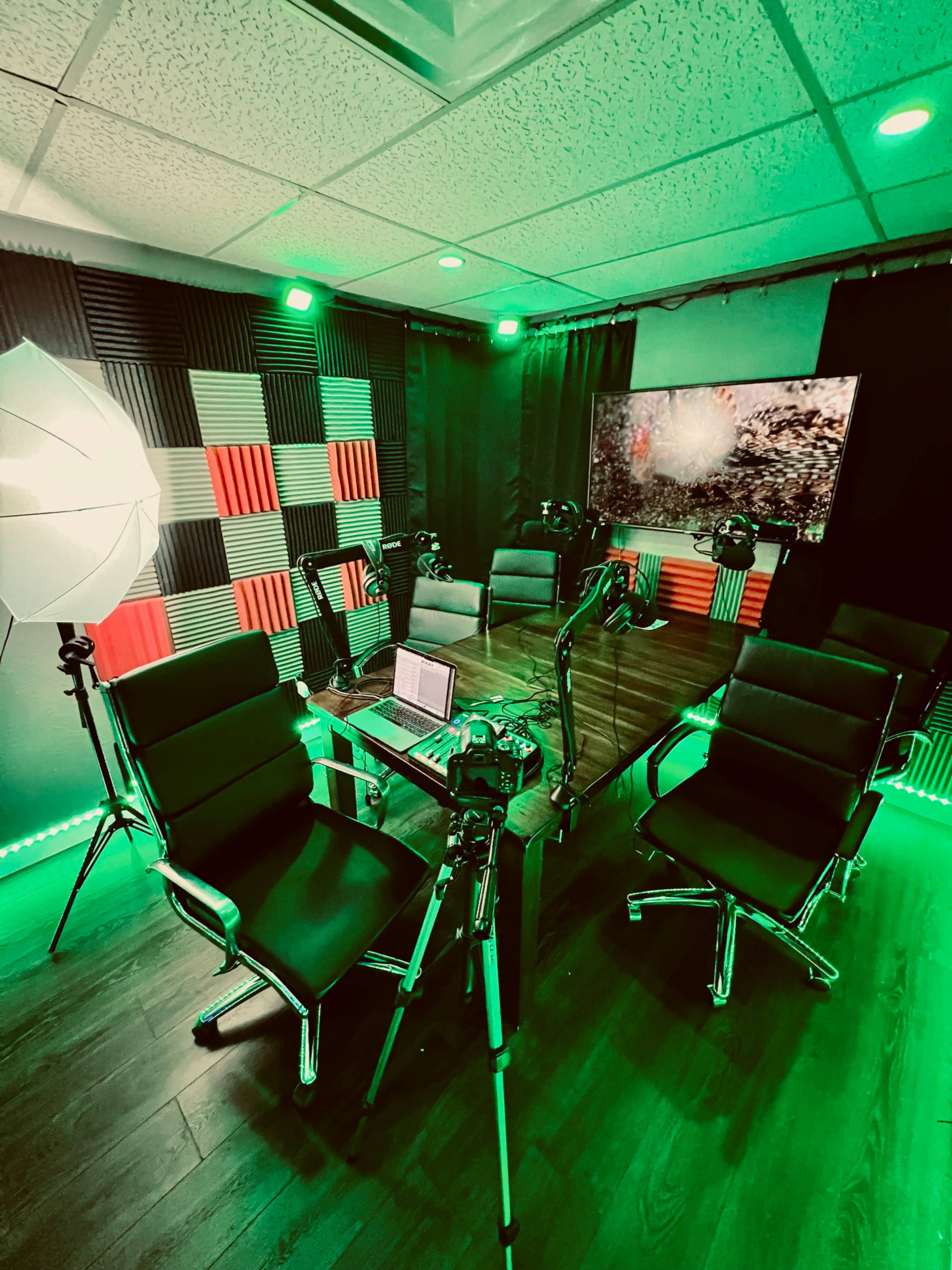 A modern studio setup with a large wooden table surrounded by black chairs, equipped with multiple cameras and lighting, and featuring green ambient lighting and a patterned backdrop.