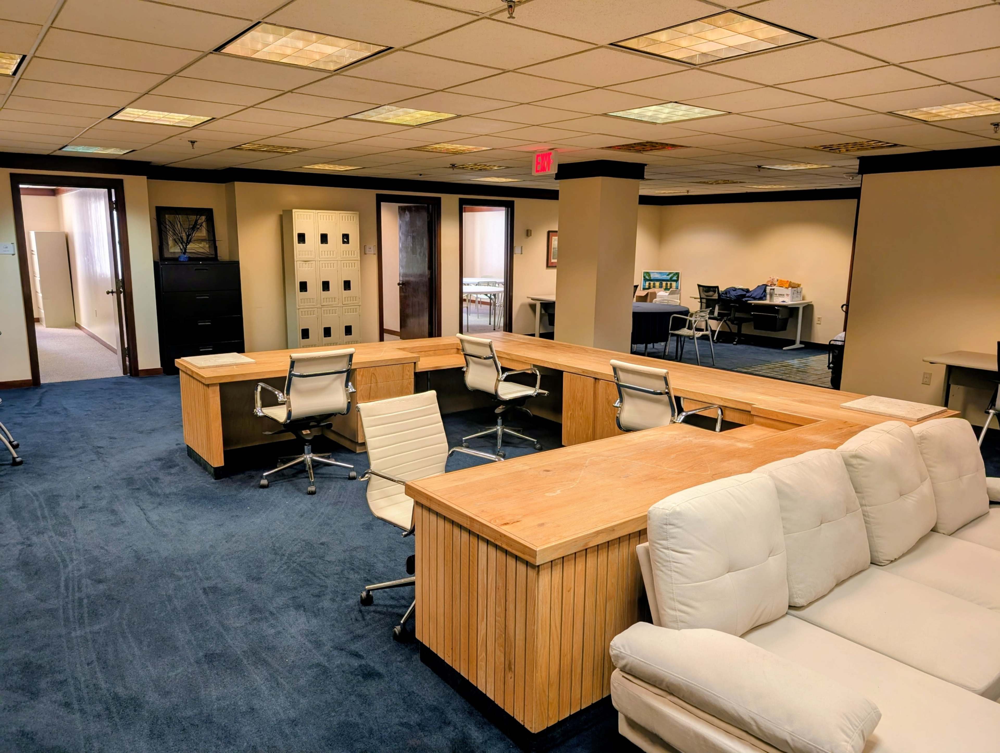 The image shows an office space featuring wooden desks, a sofa, and various office furniture in a carpeted area.