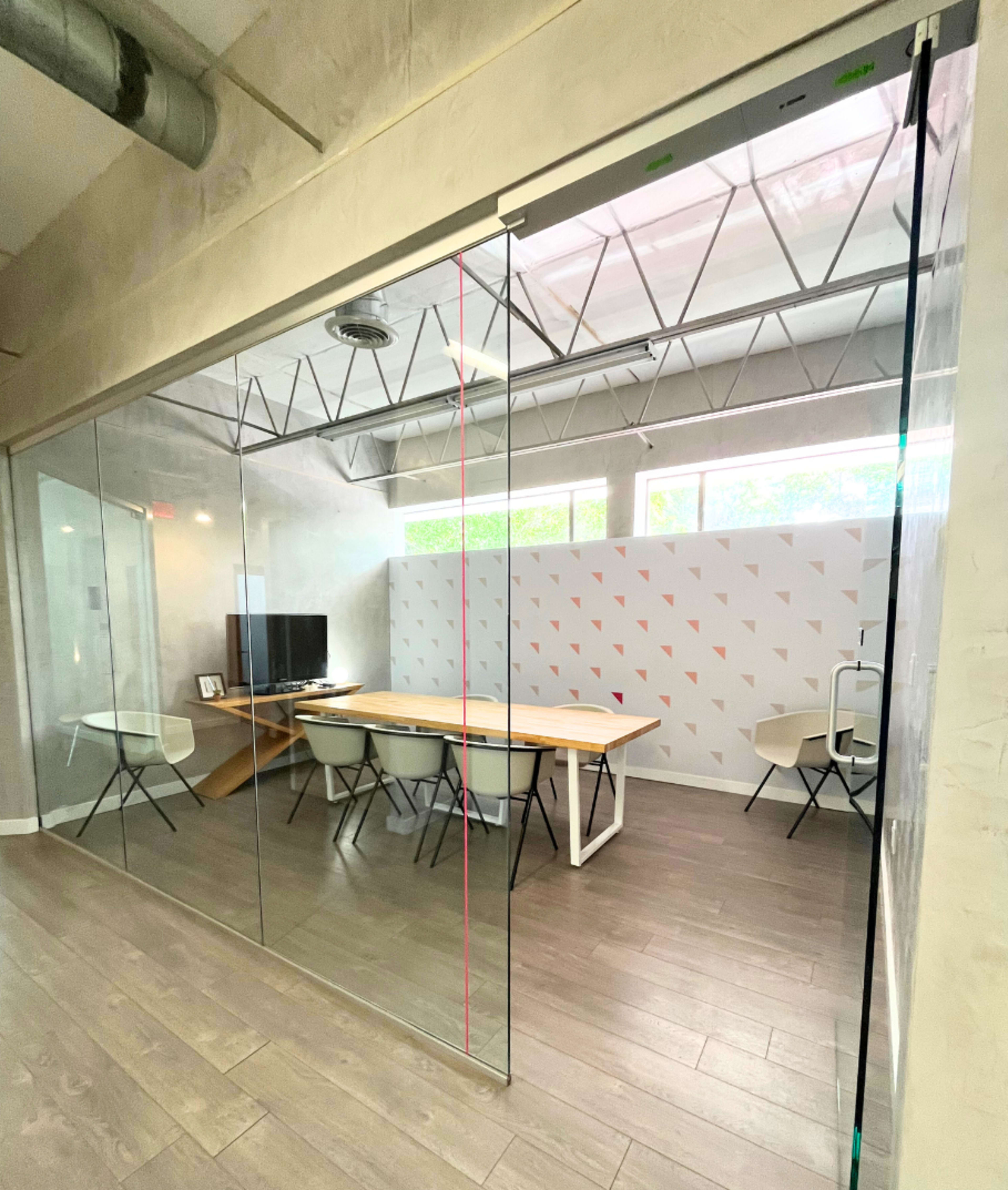 The image shows a modern conference room enclosed by glass walls, featuring a long wooden table, several chairs, and a television on one side.