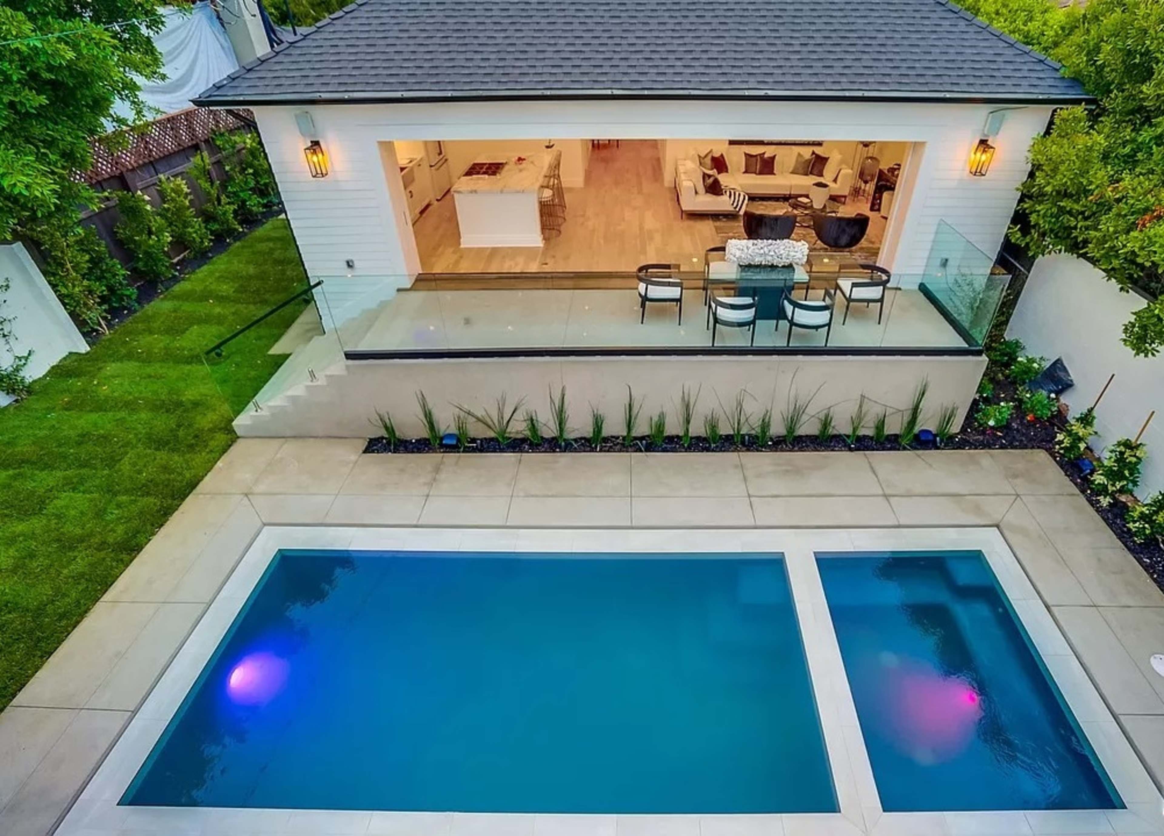 A modern house with a glass-walled balcony overlooking a rectangular swimming pool, surrounded by landscaped gardens and outdoor seating.