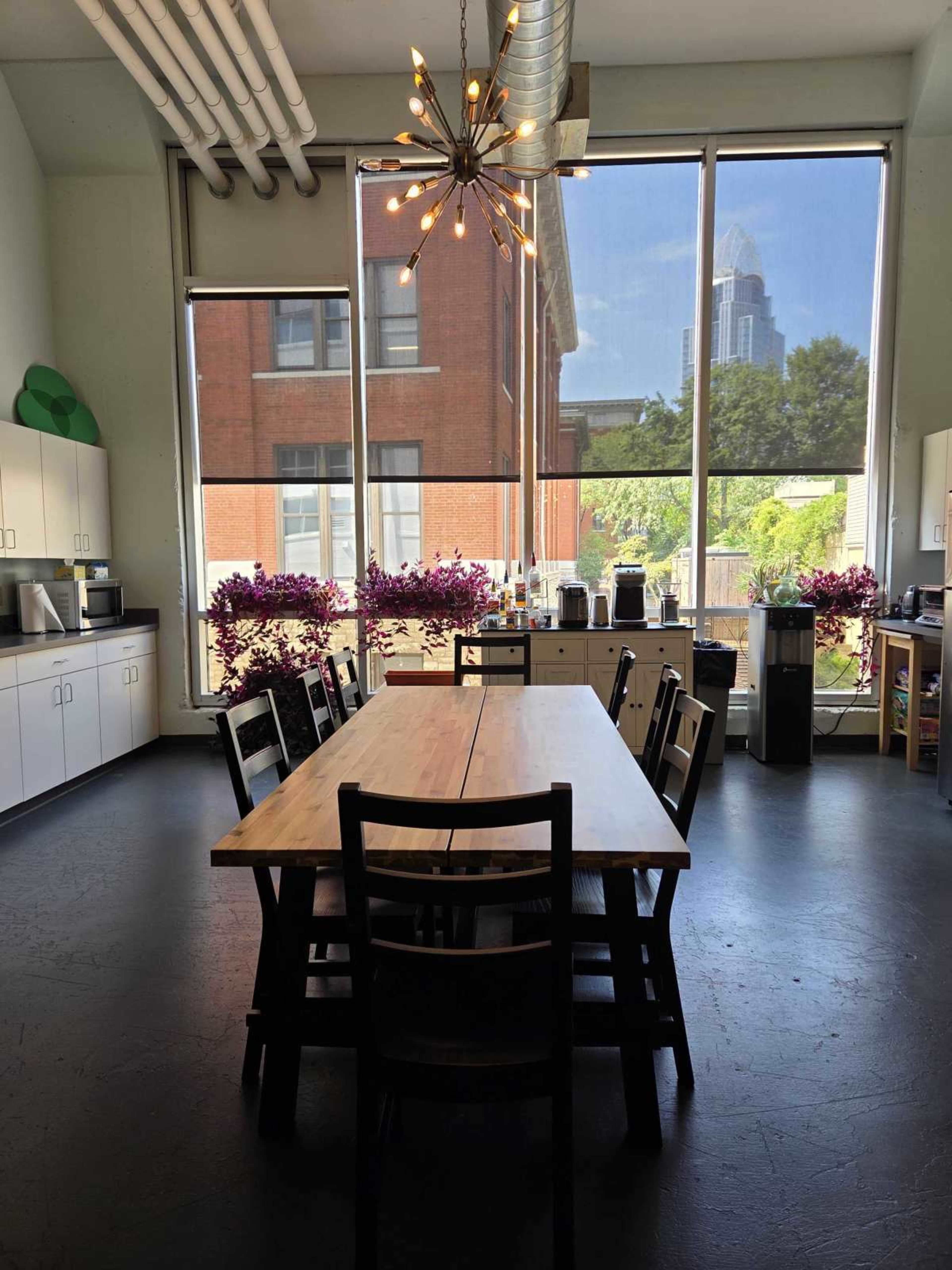 A large wooden table with chairs surrounds it in a bright kitchen area, featuring large windows that overlook a cityscape and outdoor plants.