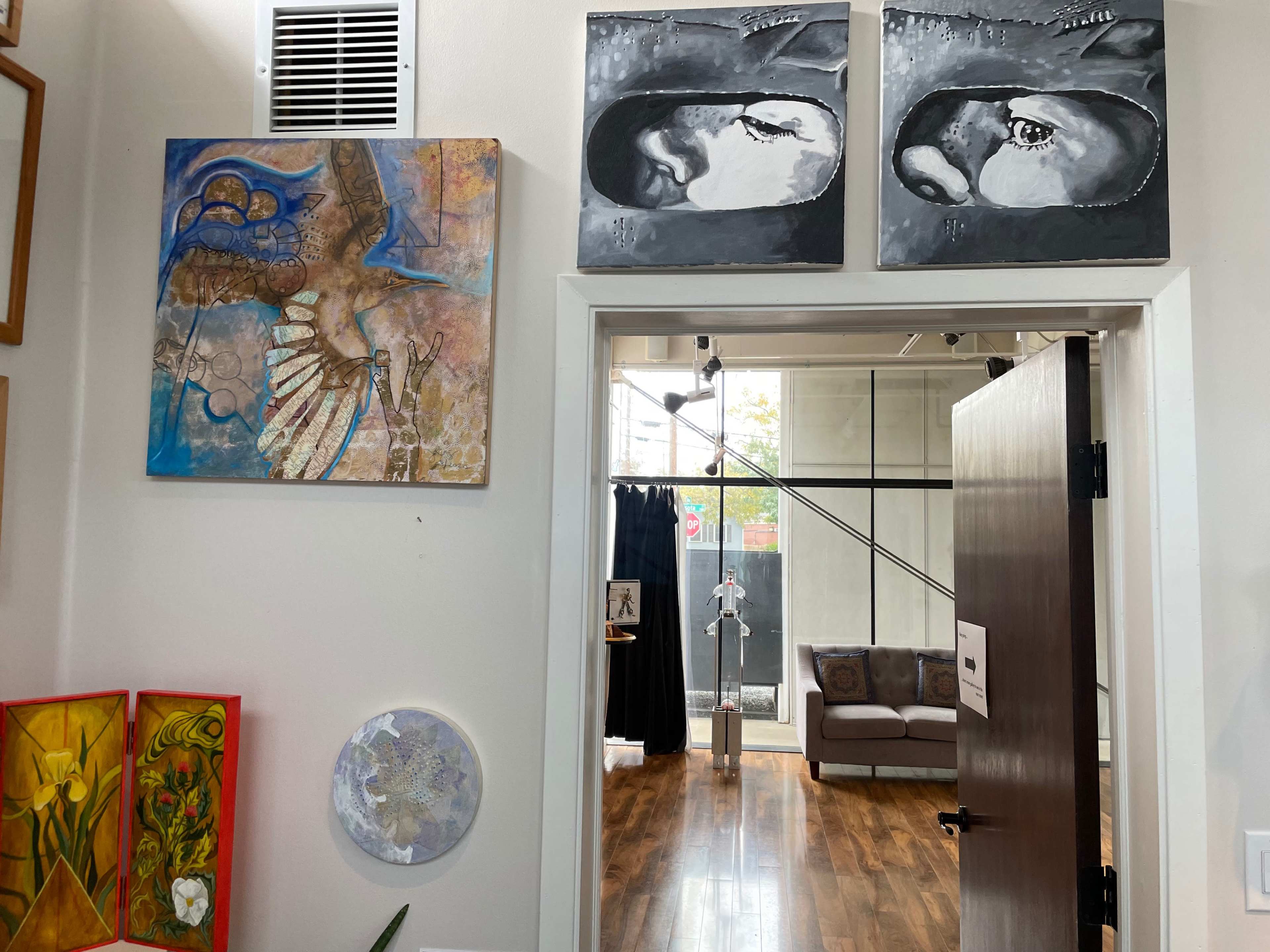 The image shows an interior space featuring artwork on the walls, including two monochrome paintings above a doorway and colorful art pieces to the left, with a modern sofa visible in the background.