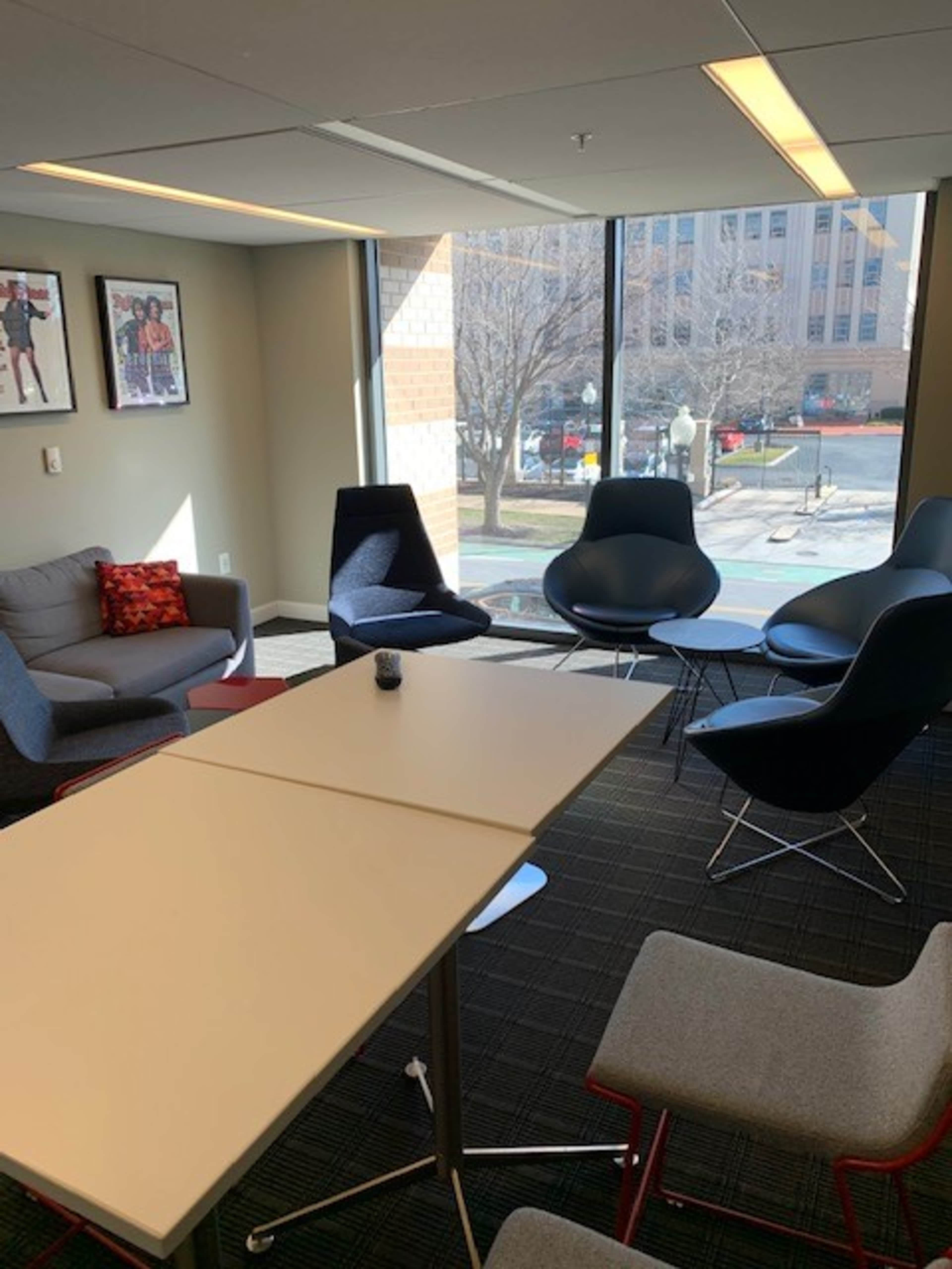 A modern office lounge area with a table and various chairs, bathed in natural light from large windows overlooking an outdoor space.