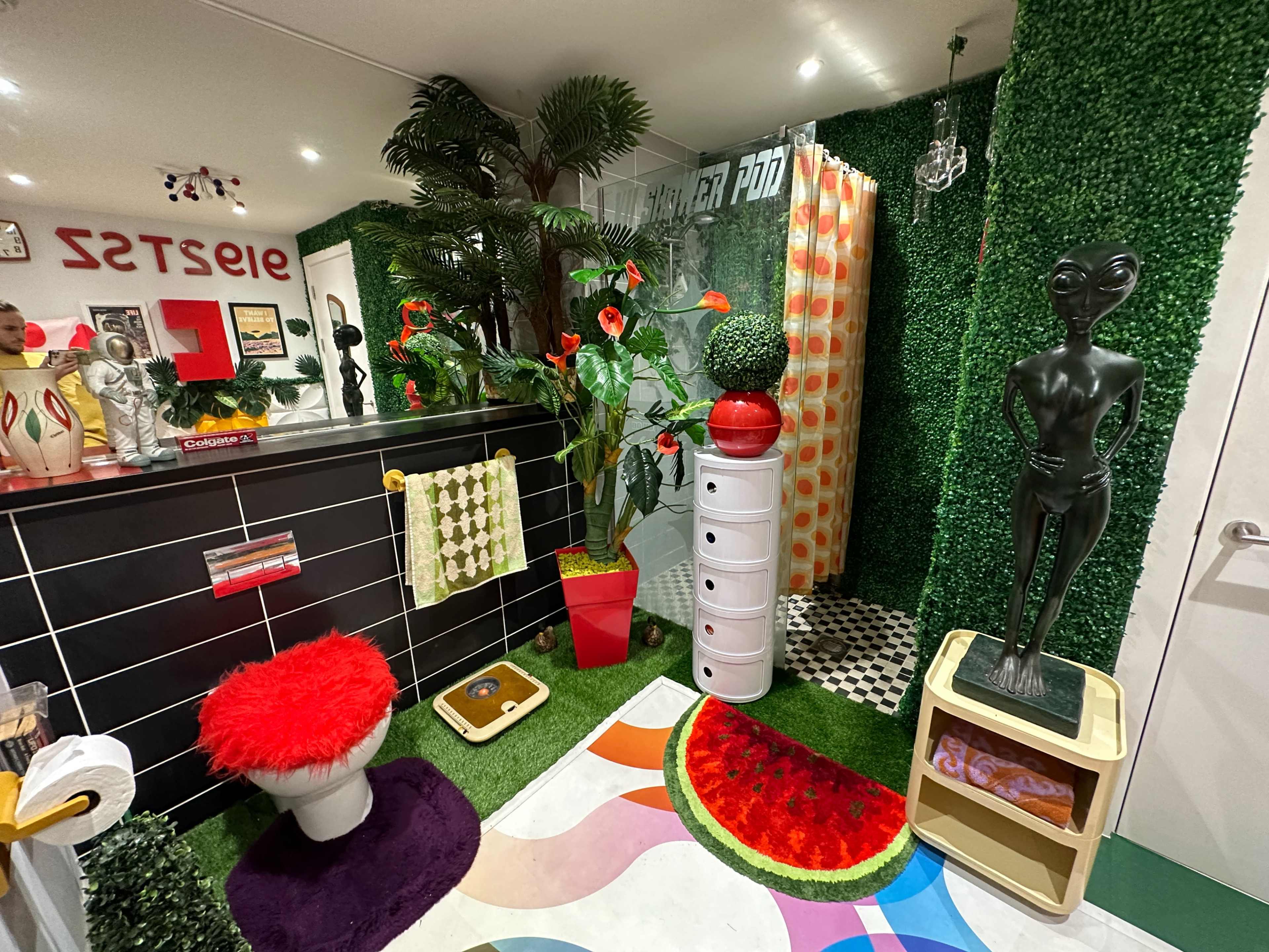 A colorful room with artificial grass walls, decorative plants, a black-and-white checkered floor, and a variety of quirky furnishings and accessories.