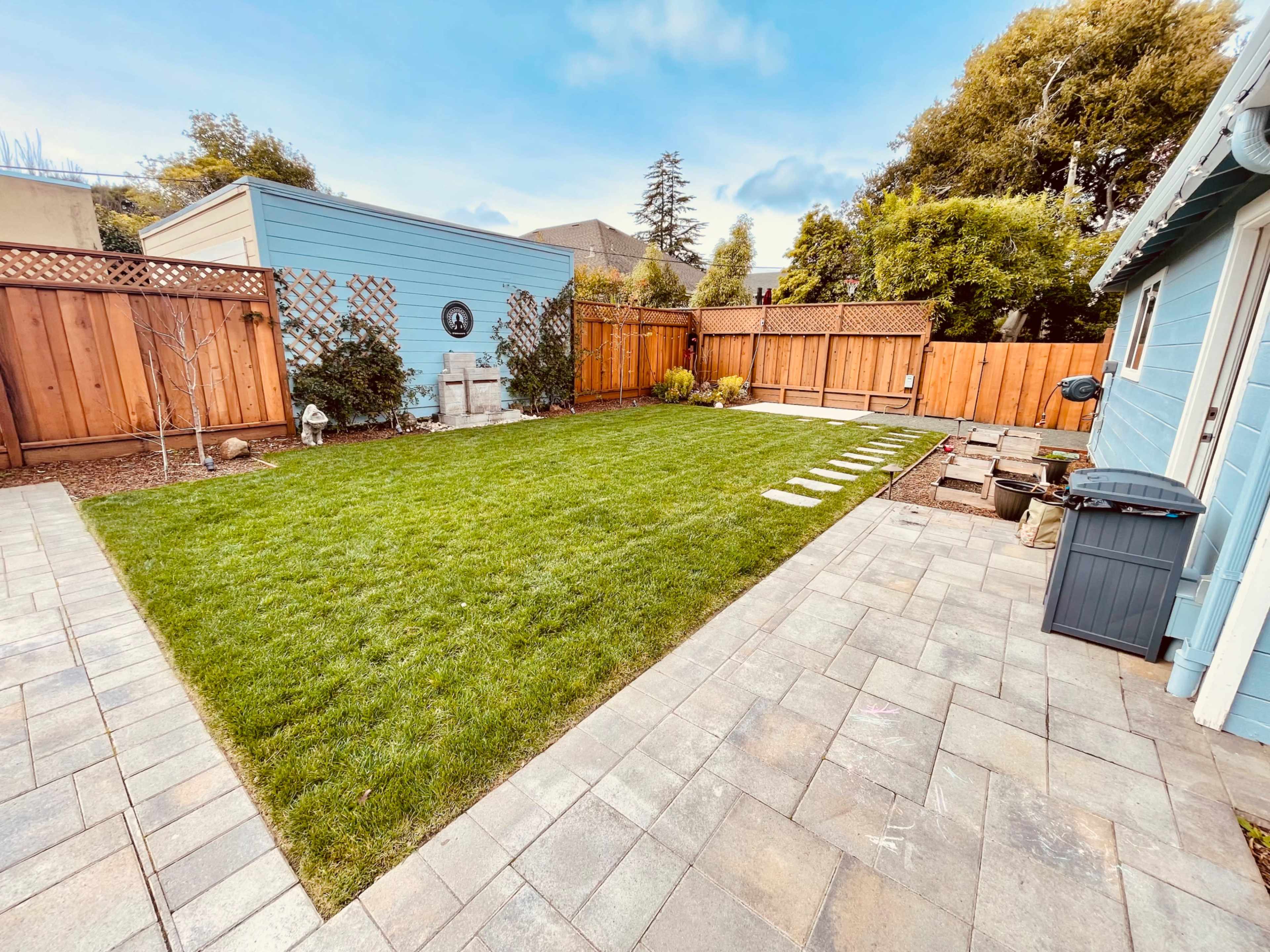 A well-maintained backyard with a green lawn, stone pathway, paved patio areas, and wooden fencing.