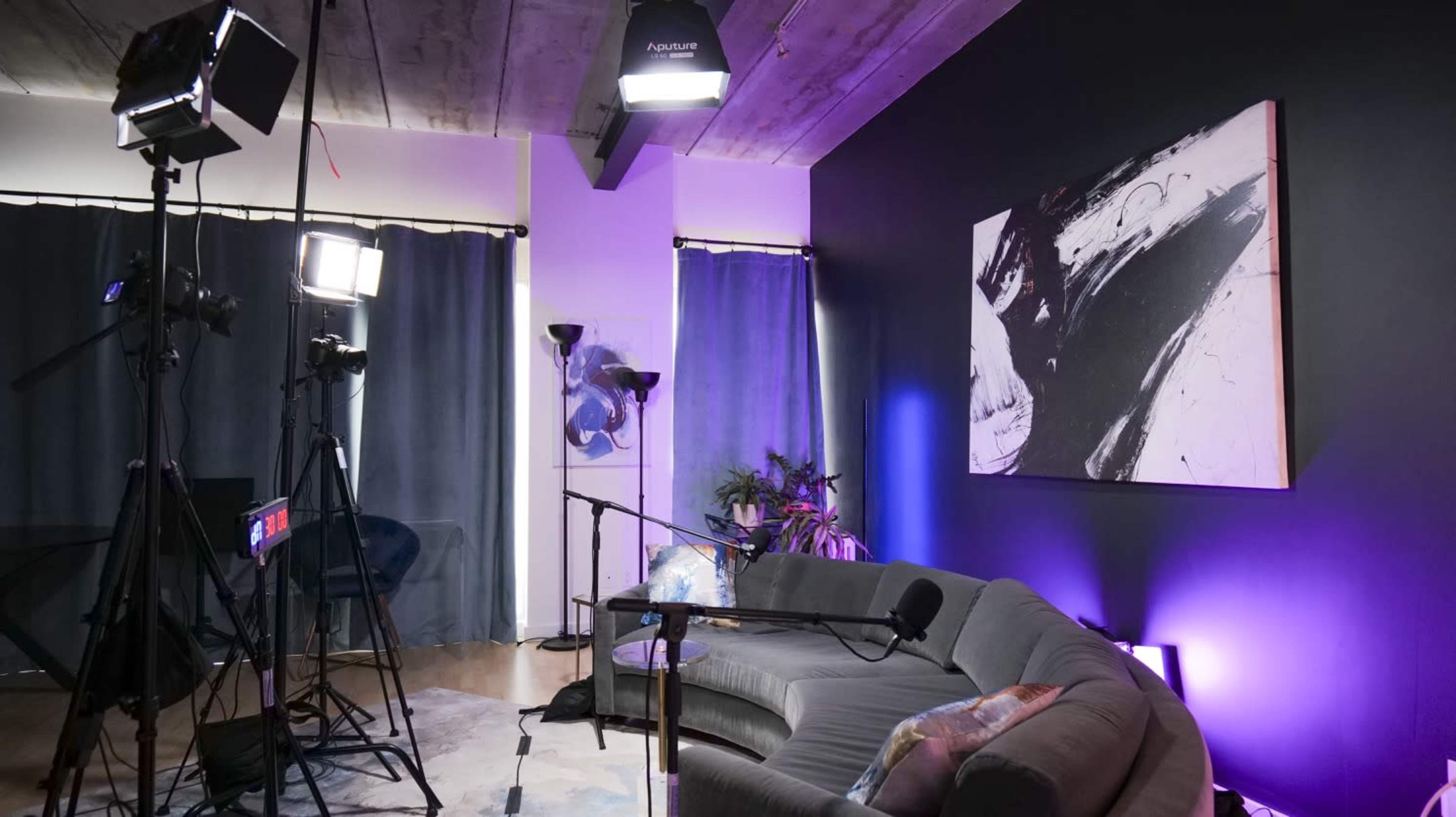 A filming setup is arranged in a living space featuring a couch, lighting equipment, and a large abstract painting on the wall.