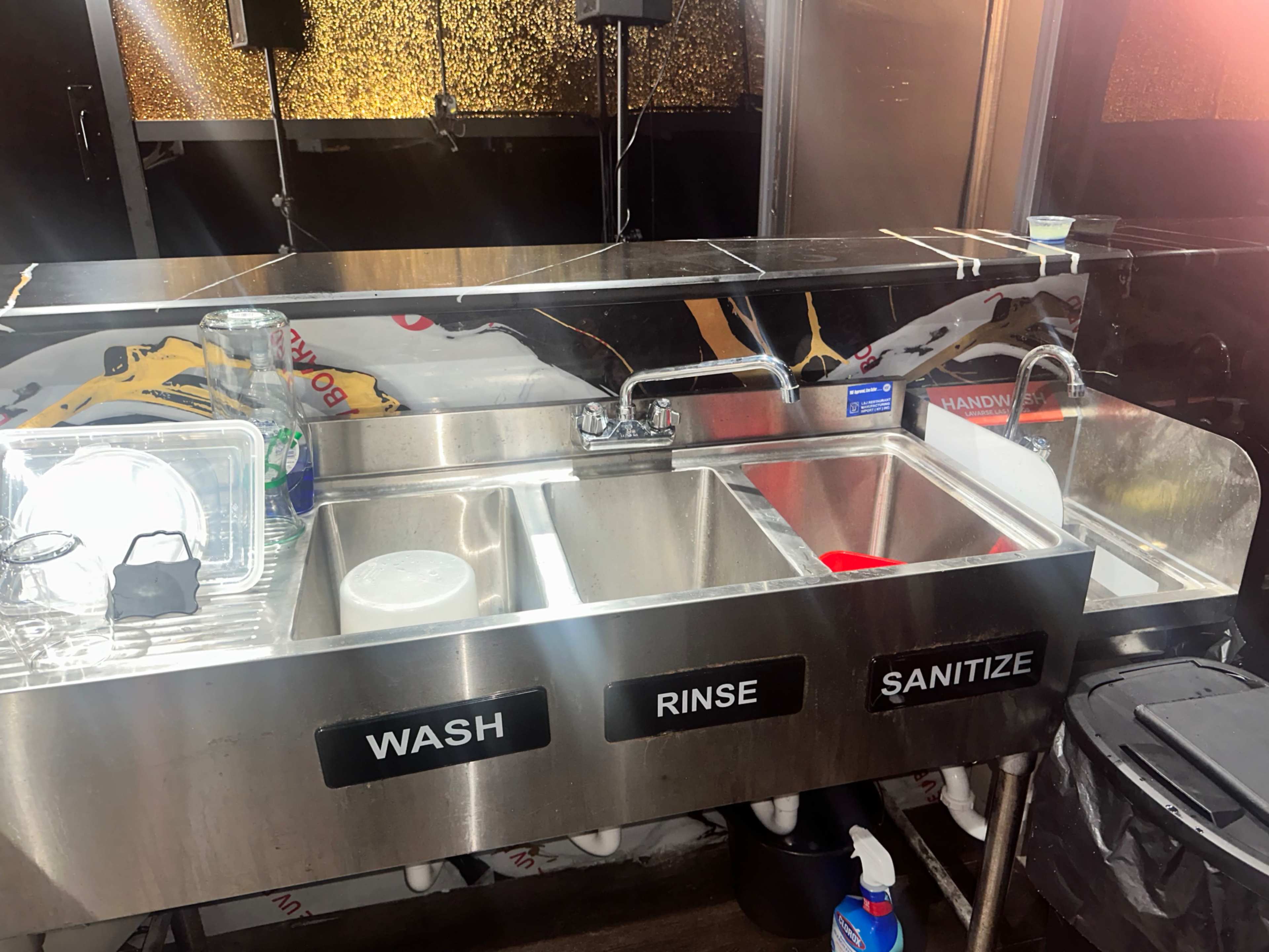 The image shows a three-compartment sink labeled "WASH," "RINSE," and "SANITIZE," with various cleaning supplies and dishes beside it.