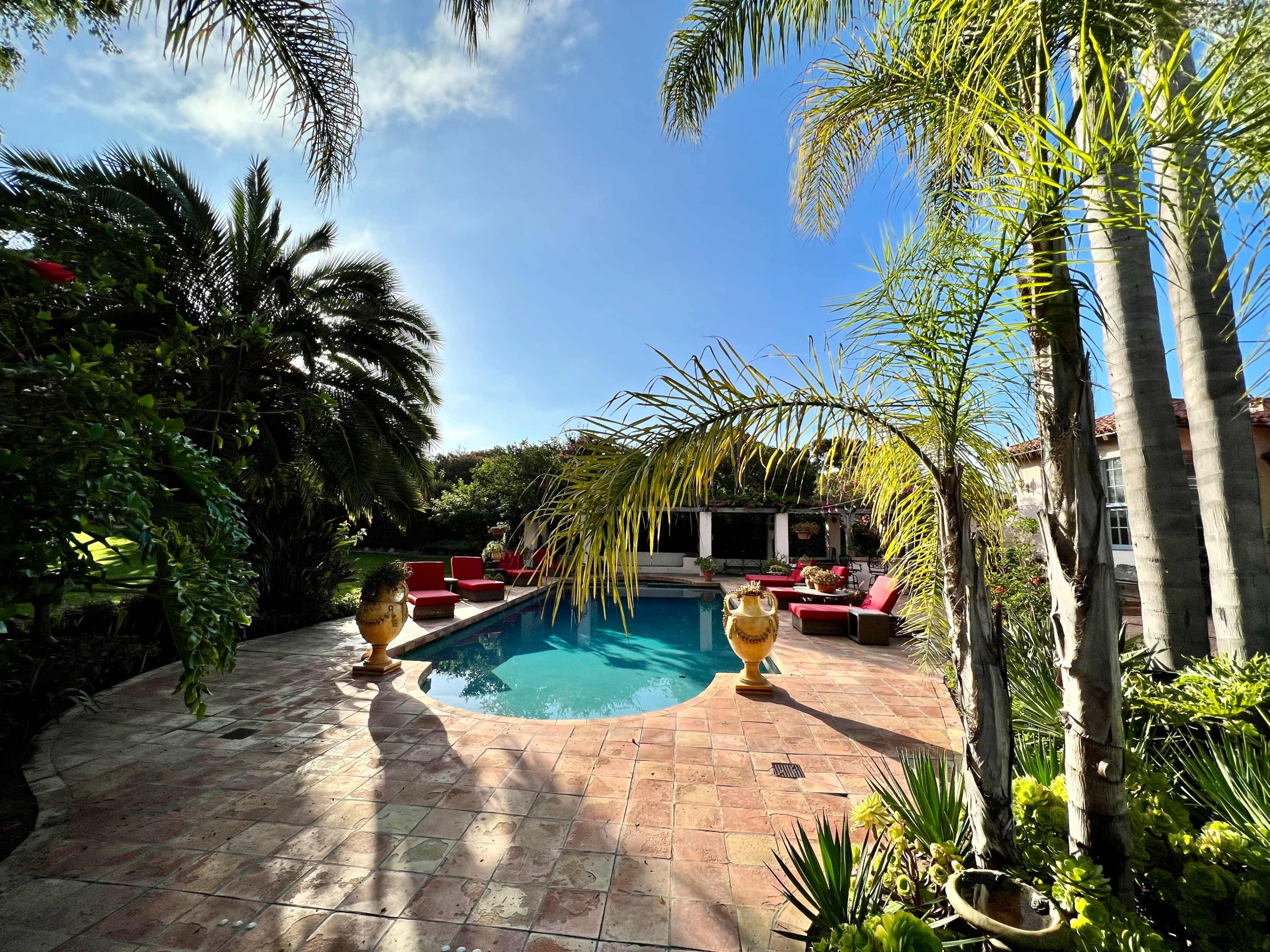 A swimming pool surrounded by palm trees and red lounge chairs is set in a landscaped backyard.