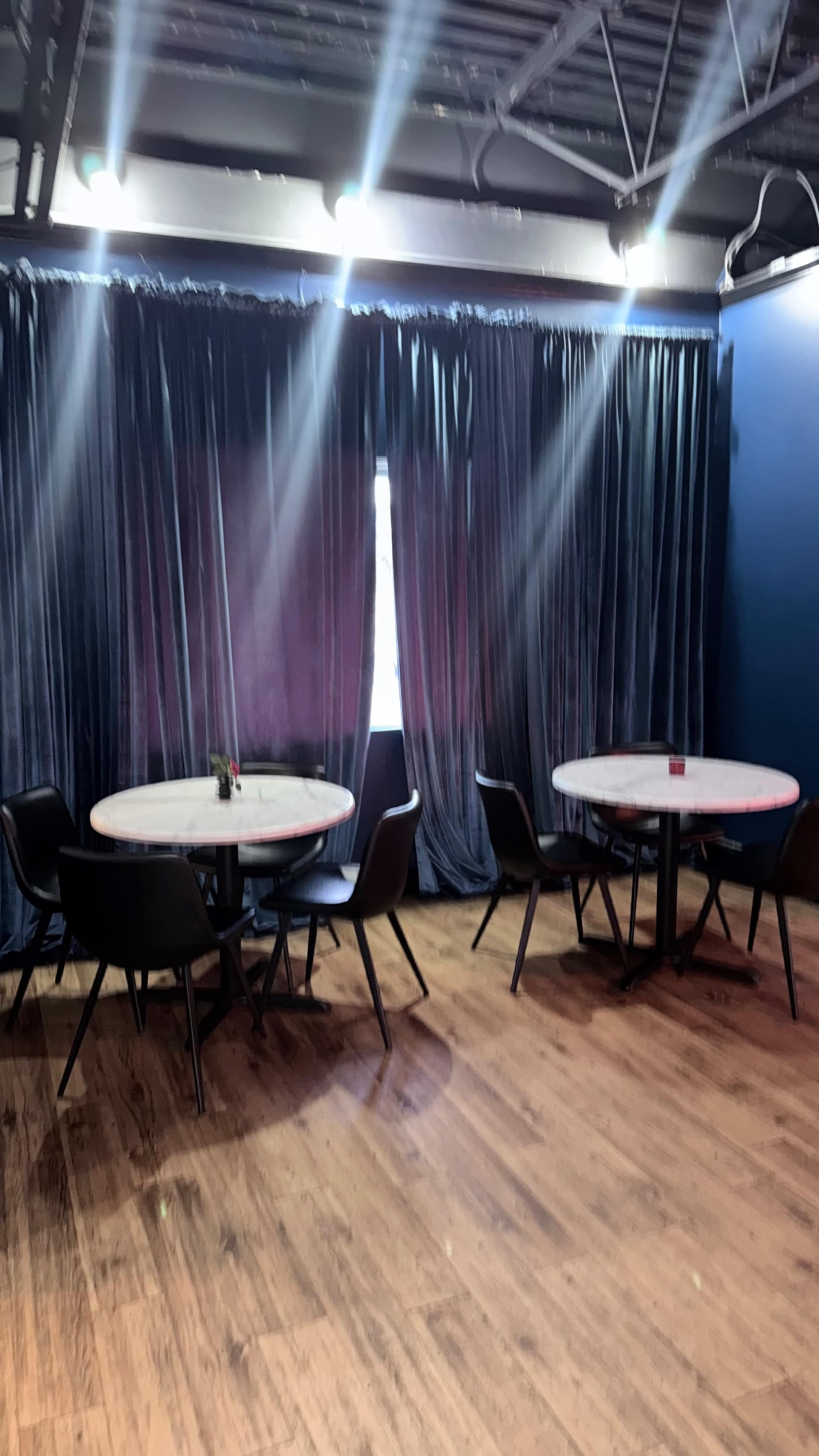 The image shows a modern interior of a dining area with several round tables and black chairs, surrounded by dark curtains and wooden flooring.