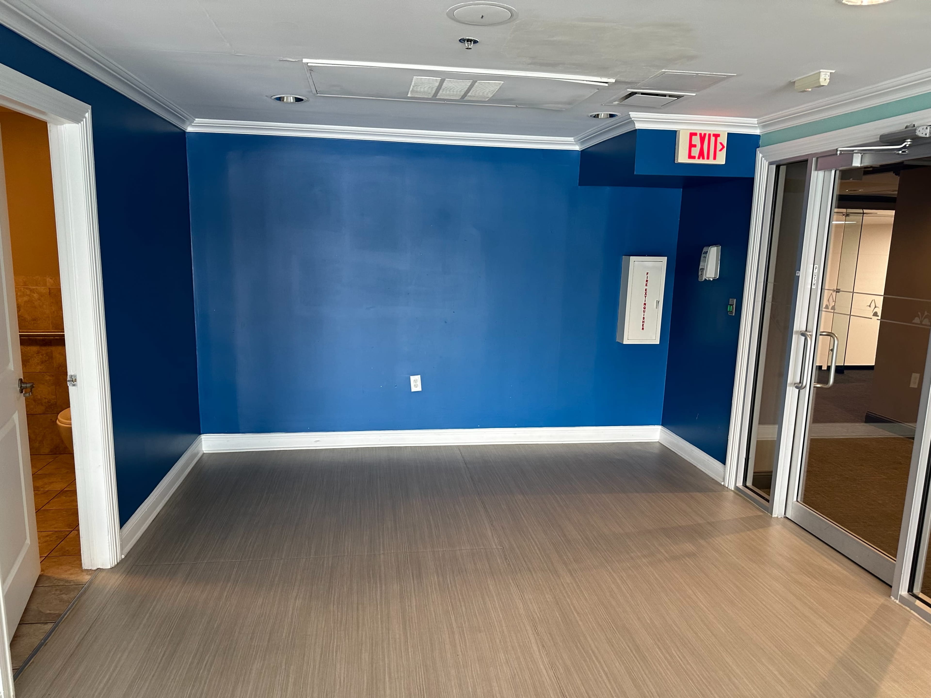 A small, empty room with a blue wall, a white exit sign, and two doorways leading to adjacent areas.