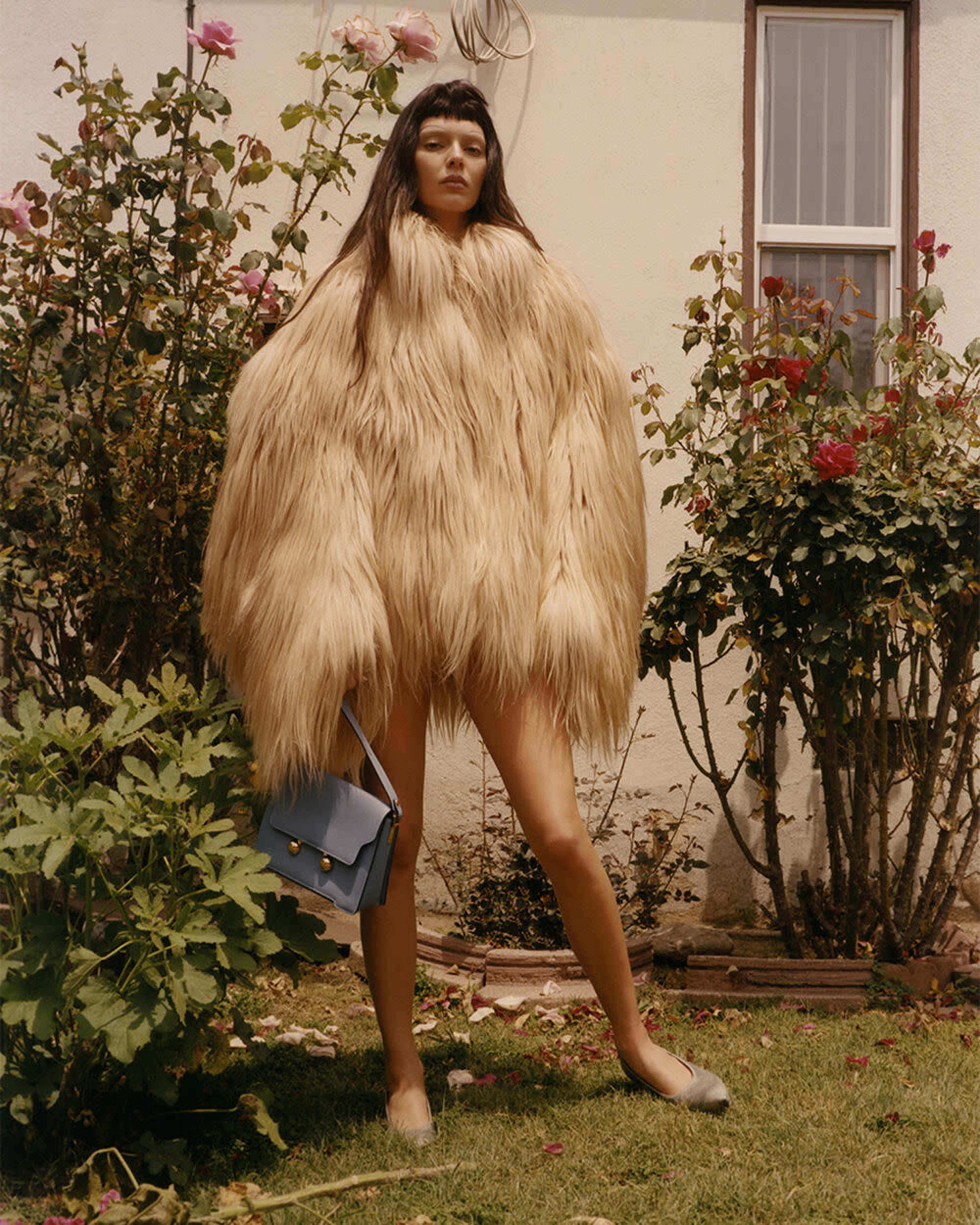A person wearing a large, fluffy coat stands in a garden surrounded by rose bushes.