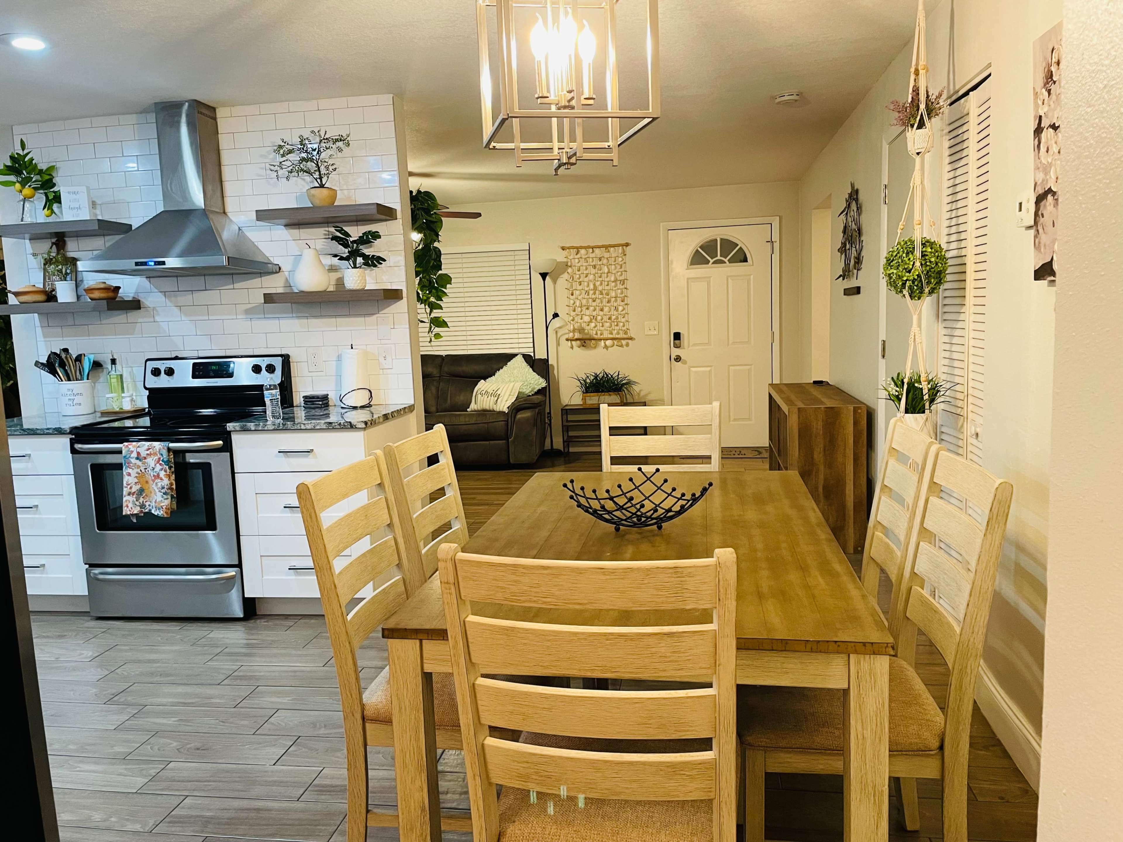 A modern kitchen and dining area features a wooden dining table with six chairs, stainless steel appliances, and decorative plants on shelves.