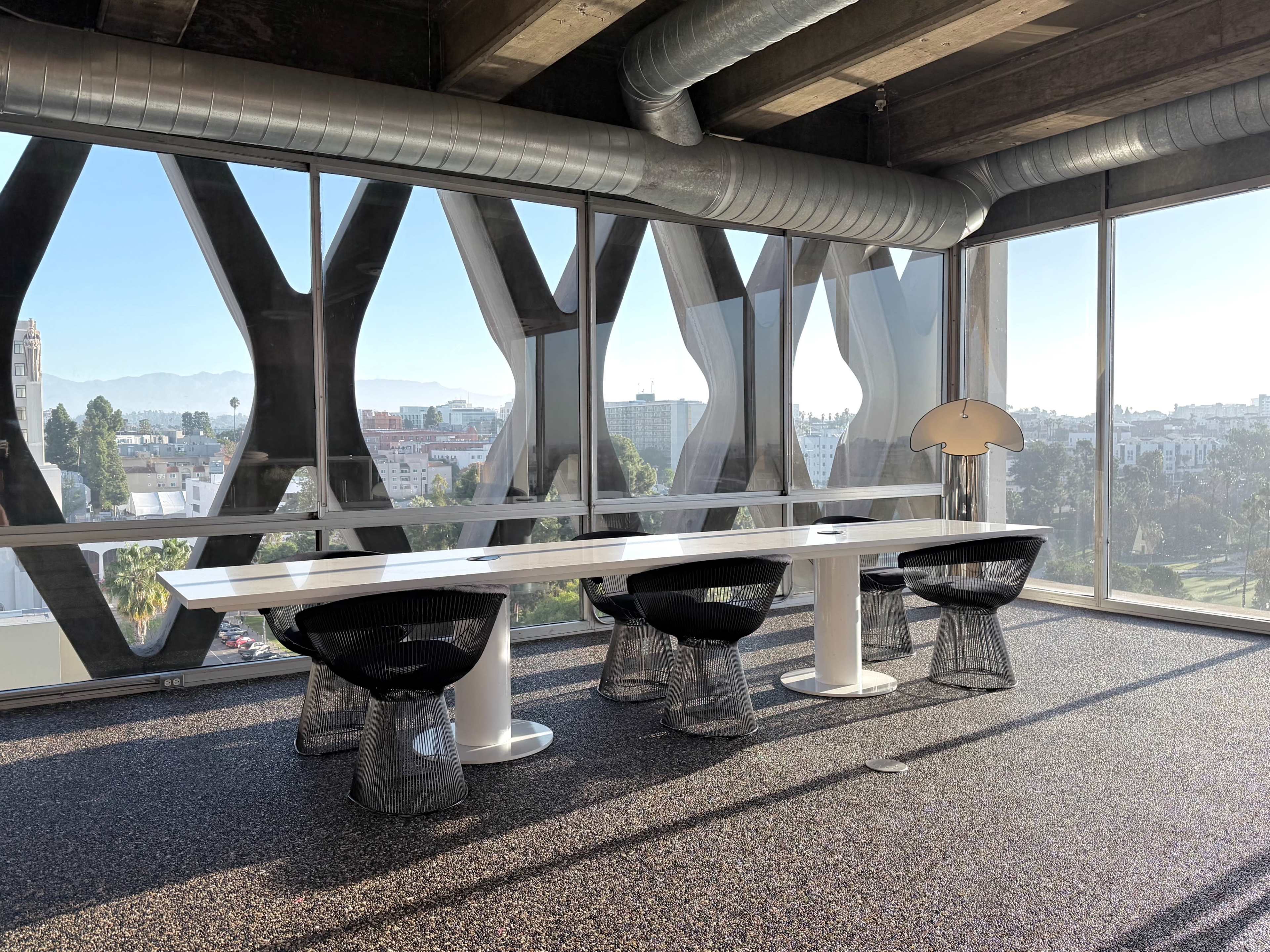 A modern office space features a long white table surrounded by black chairs, with large windows providing a view of the cityscape and mountains in the background.