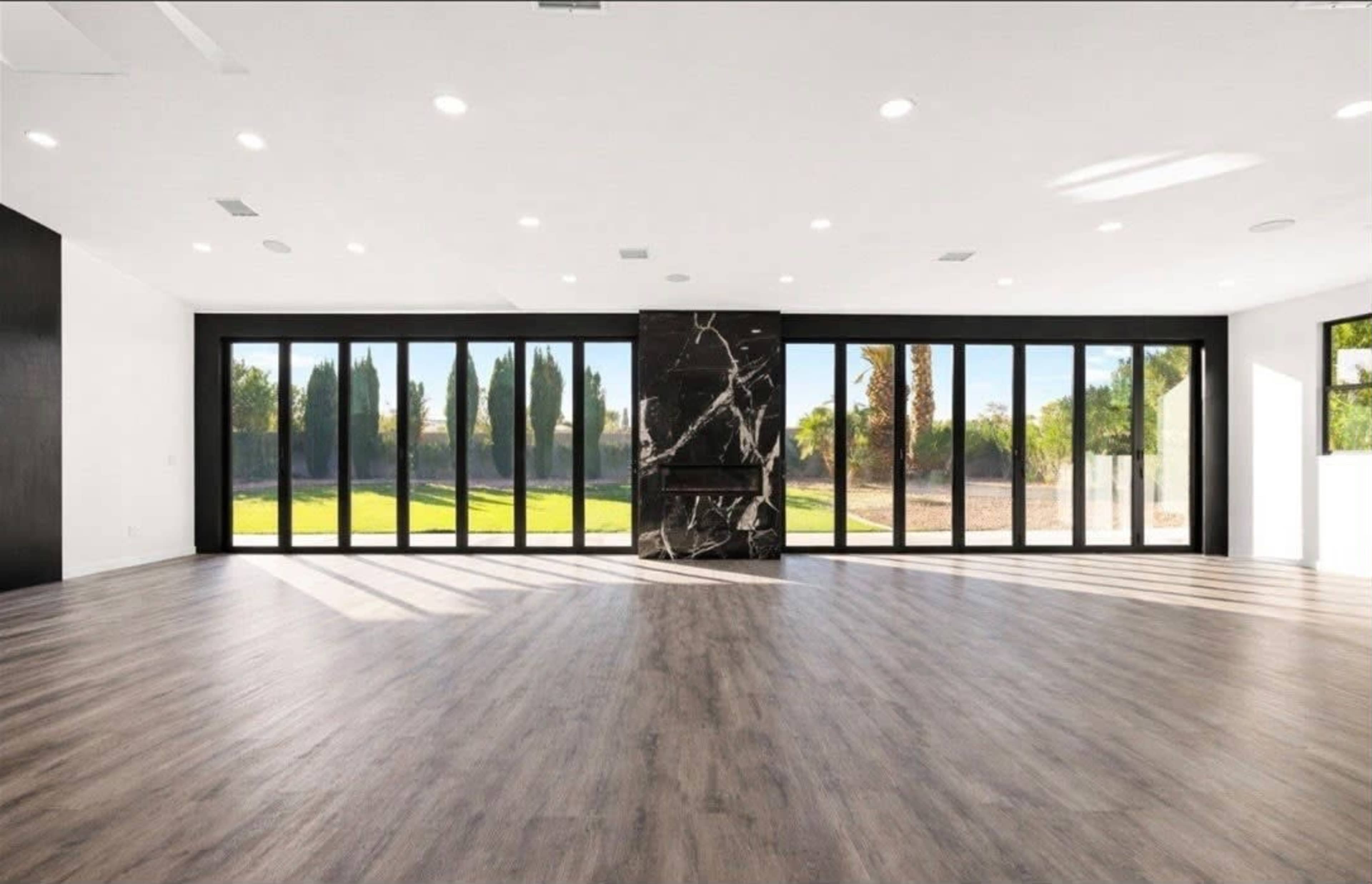 The image shows a spacious, modern interior with large glass doors facing a green landscape, featuring a black stone fireplace as a focal point.