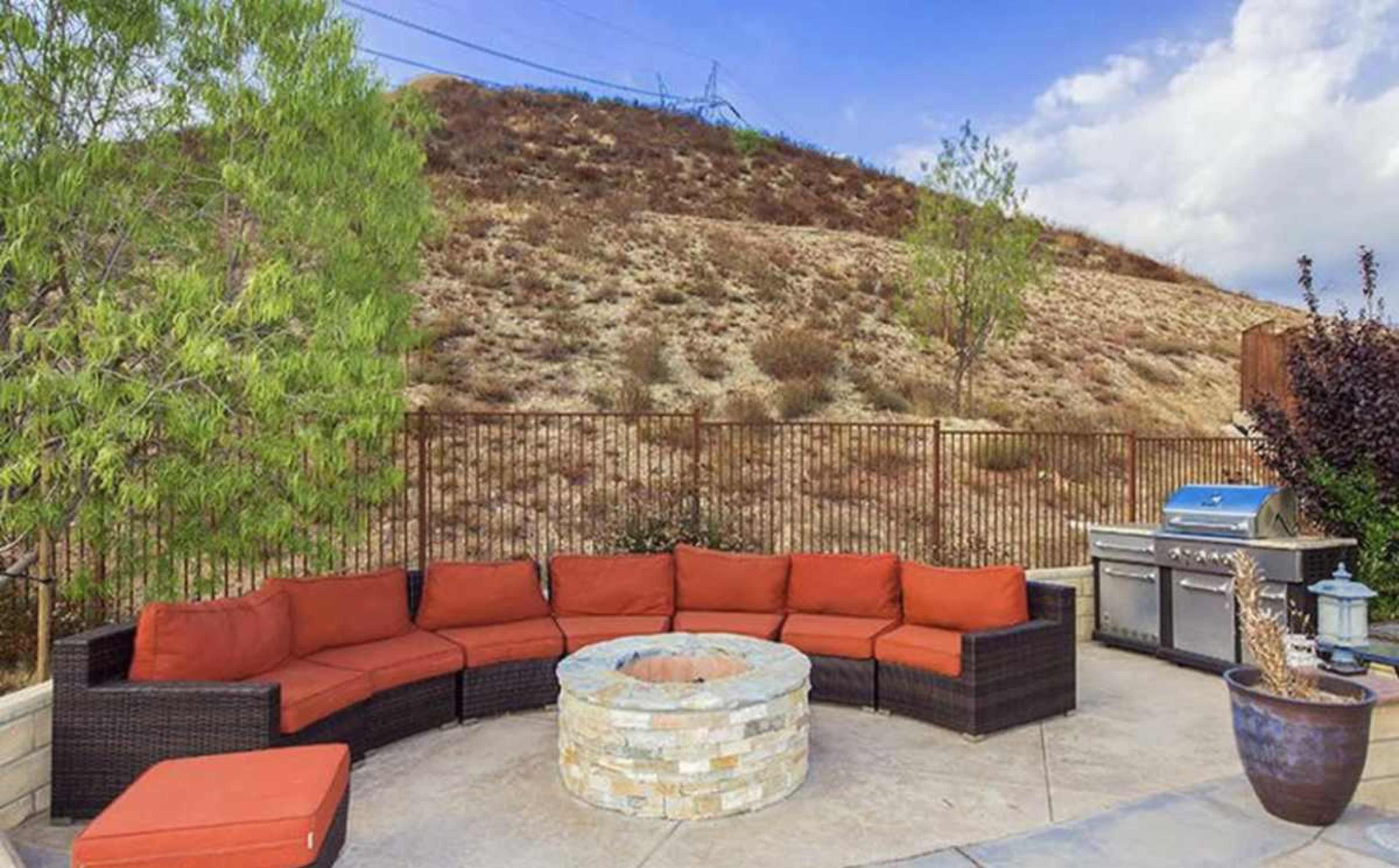The image shows an outdoor patio area with a circular stone fire pit, a curved seating arrangement covered in orange cushions, and a metal barbecue grill behind it, set against a brown hillside.