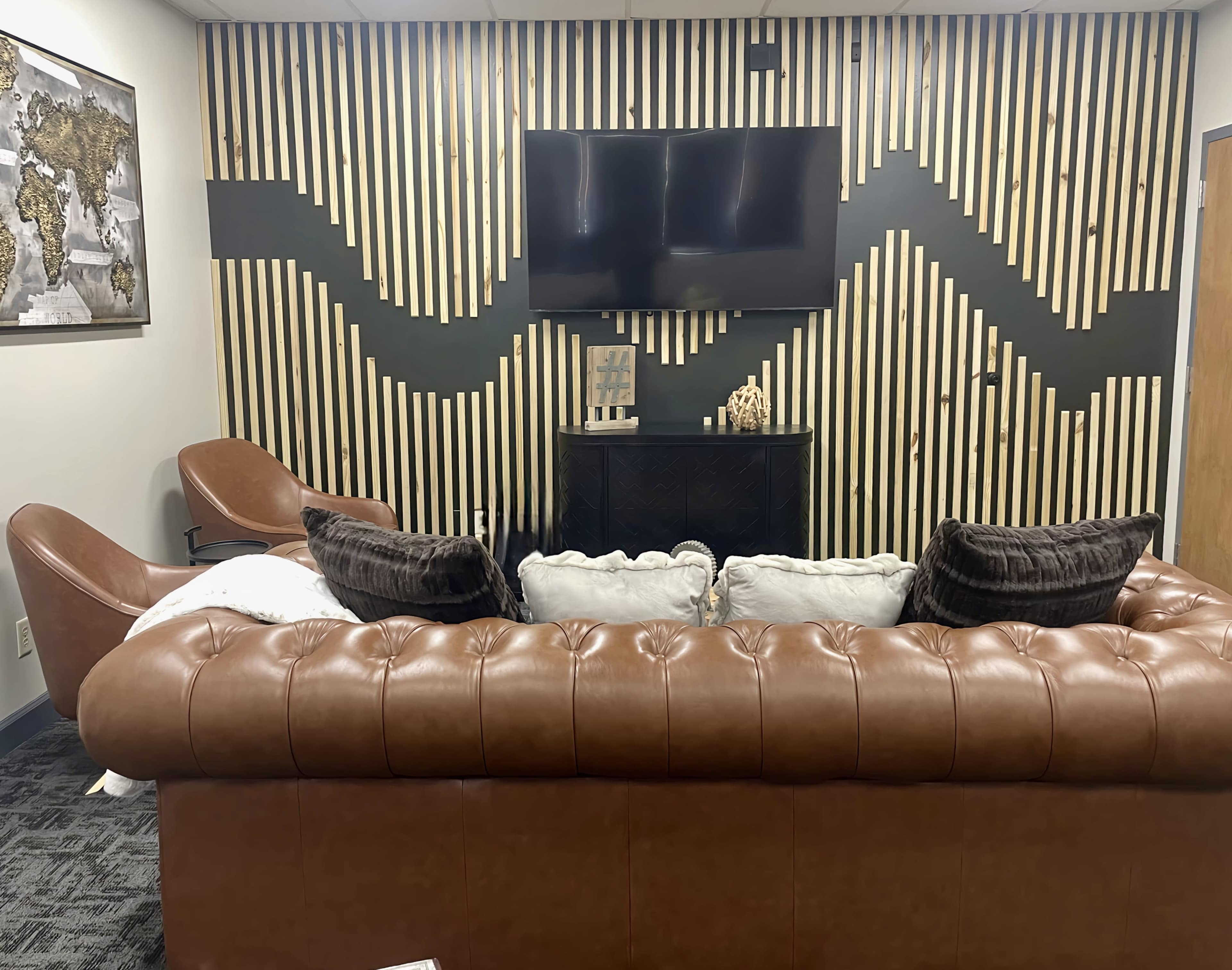 A modern lounge area with a brown leather chesterfield sofa, two brown armchairs, a television mounted on a striped black and blonde wood wall, and a decorative side table.