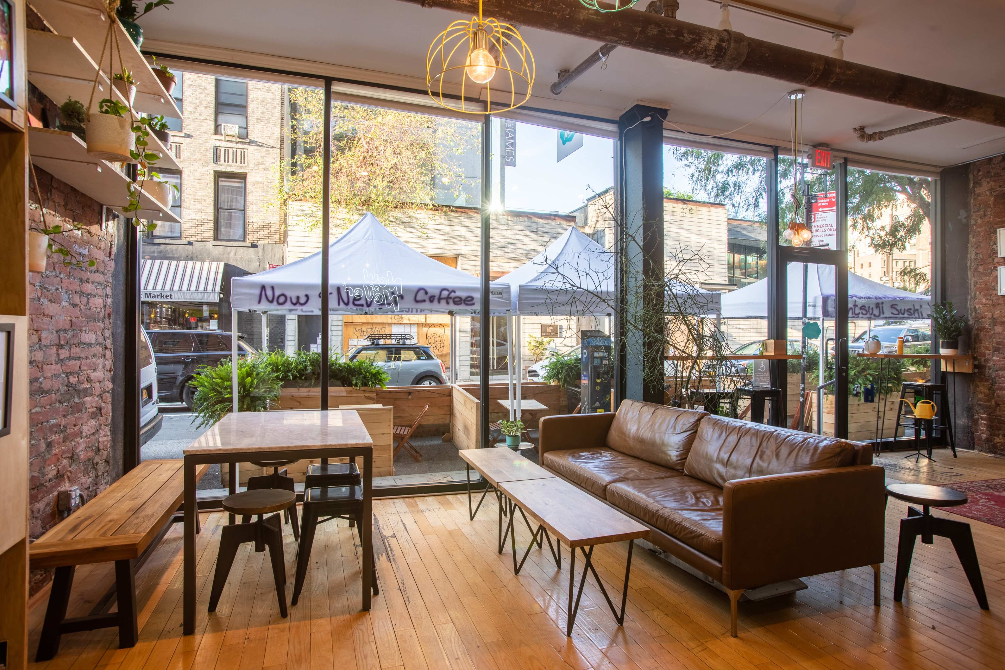 A cozy café interior with large windows overlooking an outdoor market featuring white tents, and wooden tables and a leather sofa arranged inside.