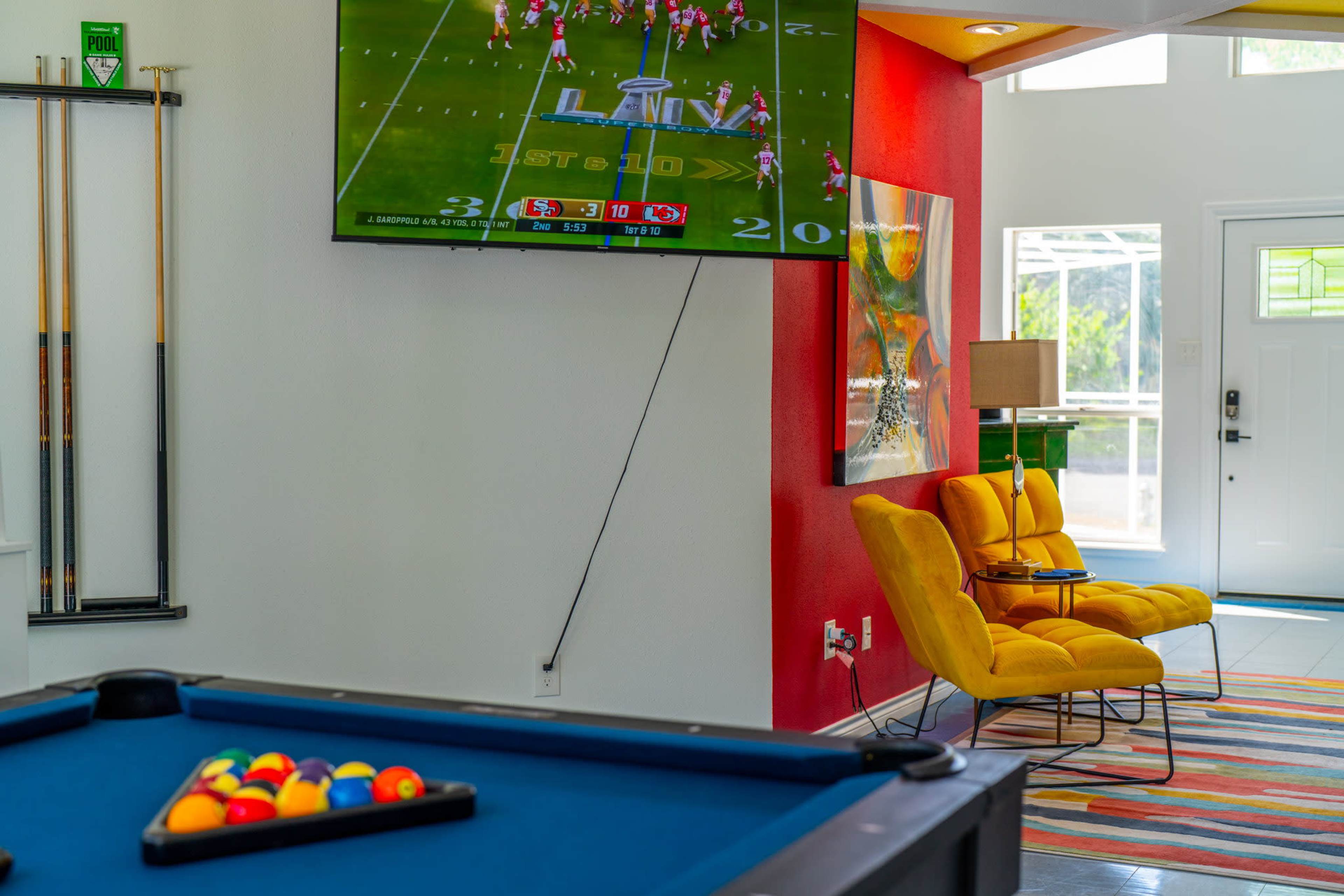 The scene features a game room with a pool table in the foreground, a television mounted on the wall displaying a football game, and a vibrant yellow chair beside a doorway.