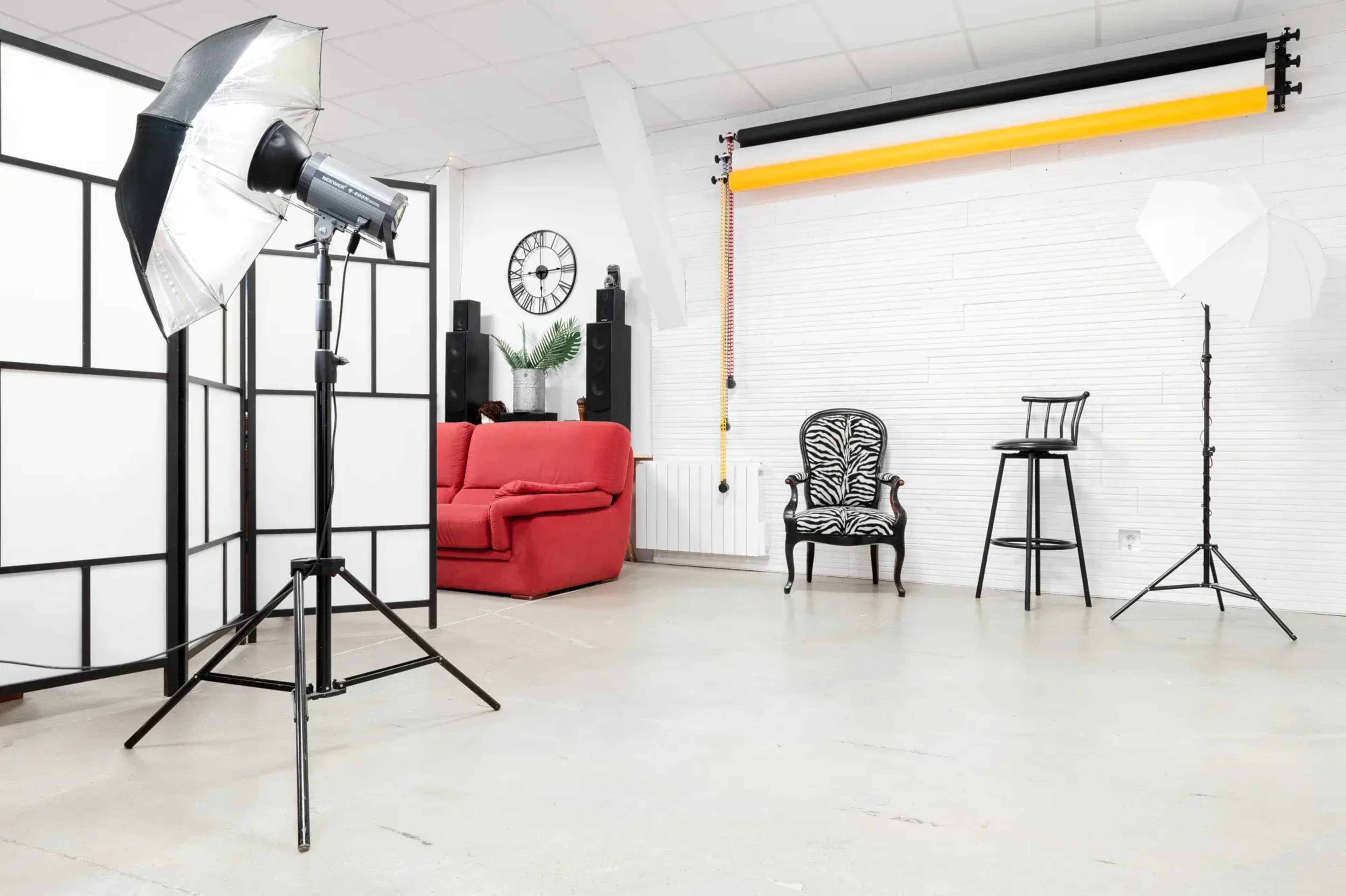 The image shows a photography studio setup featuring a red sofa, a black and white chair, various lighting equipment, and a backdrop rod with colored paper.