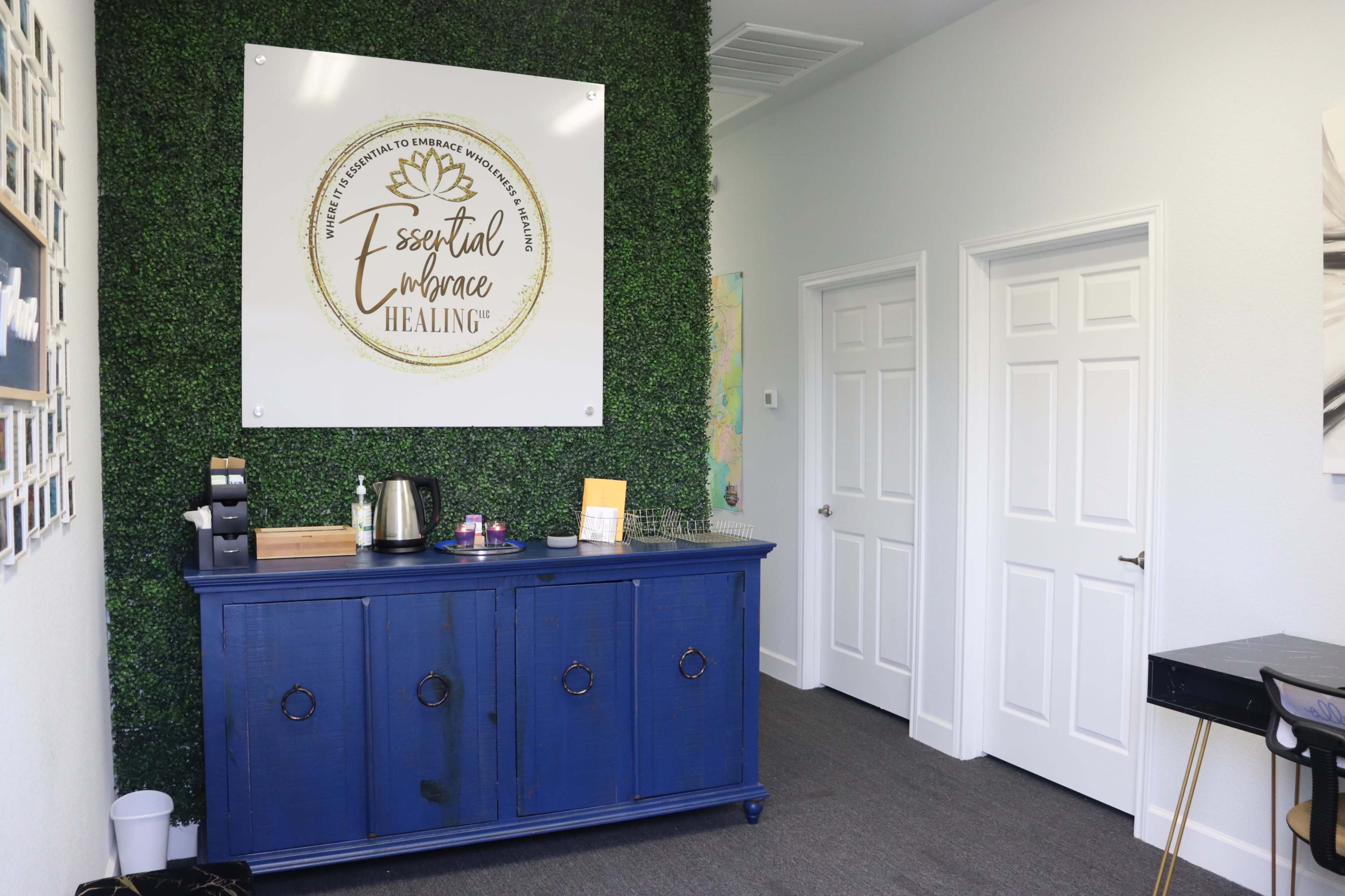 A reception area with a blue cabinet, a coffee station, and two closed white doors, all set against a green plant wall and a branded sign on the wall.