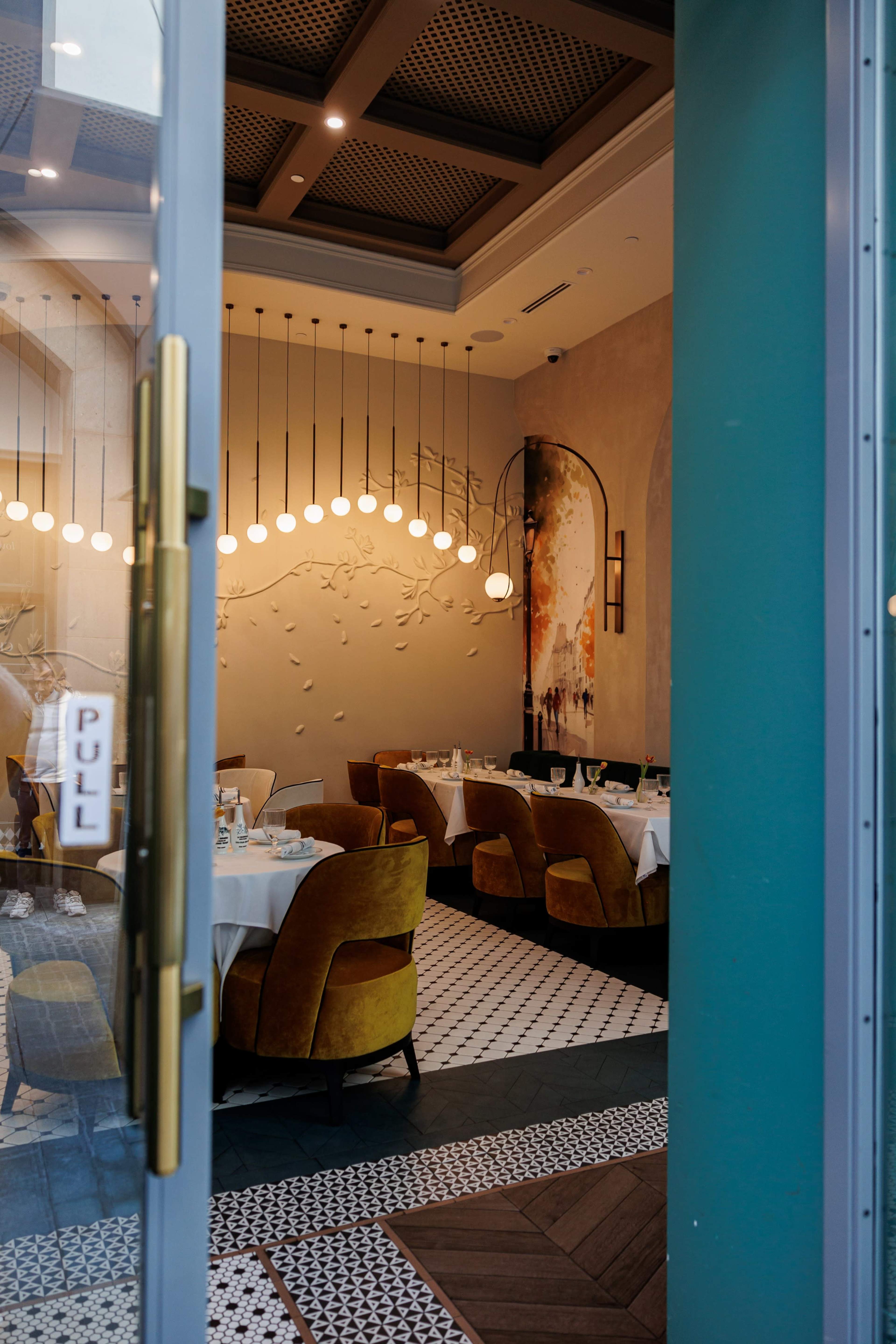 The image shows the entrance of a restaurant featuring a dining area with round tables, yellow upholstered chairs, and decorative lighting.