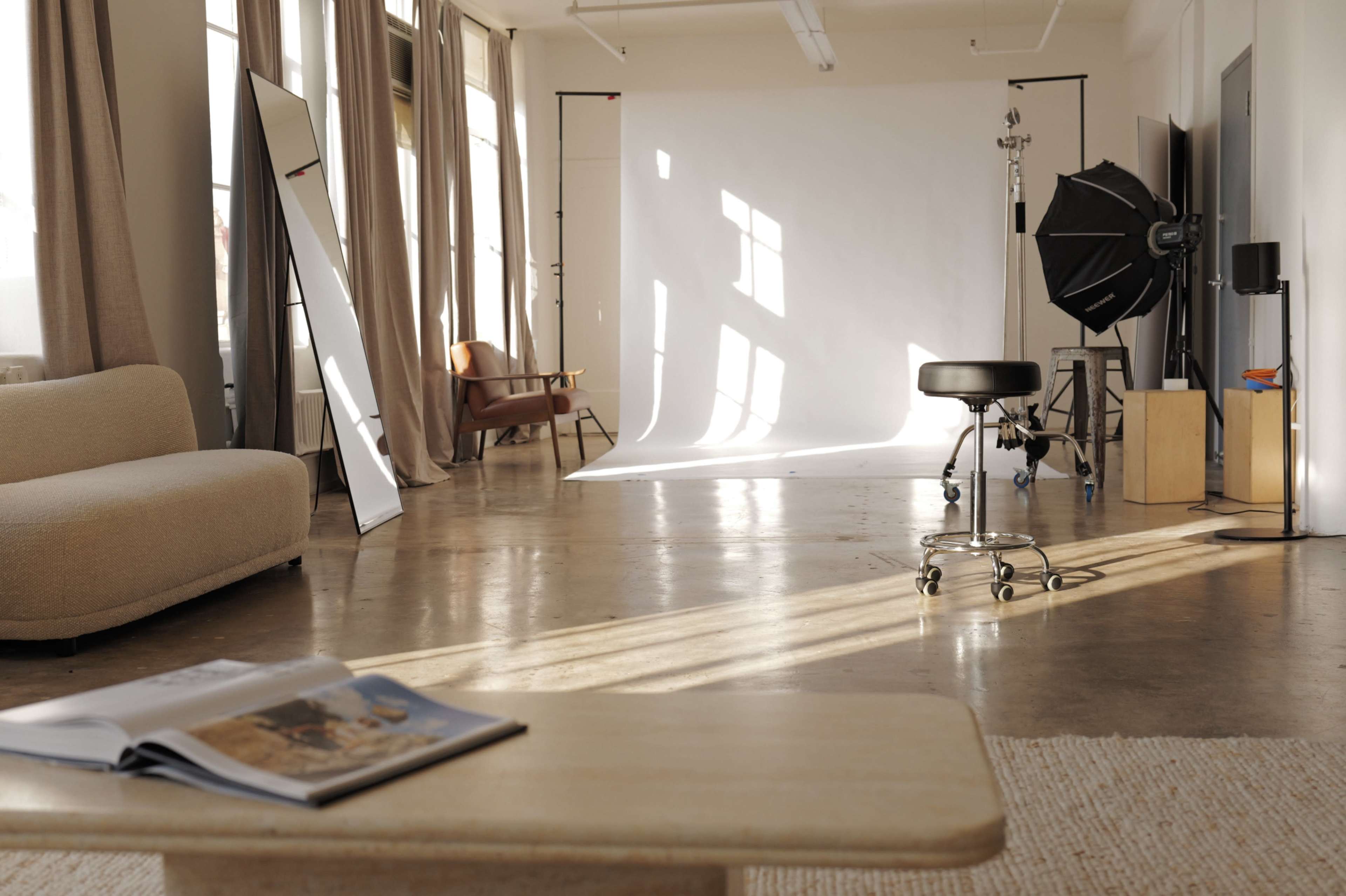 The image shows a photography studio with a white backdrop, a large softbox, a stool, and a coffee table with an open magazine.