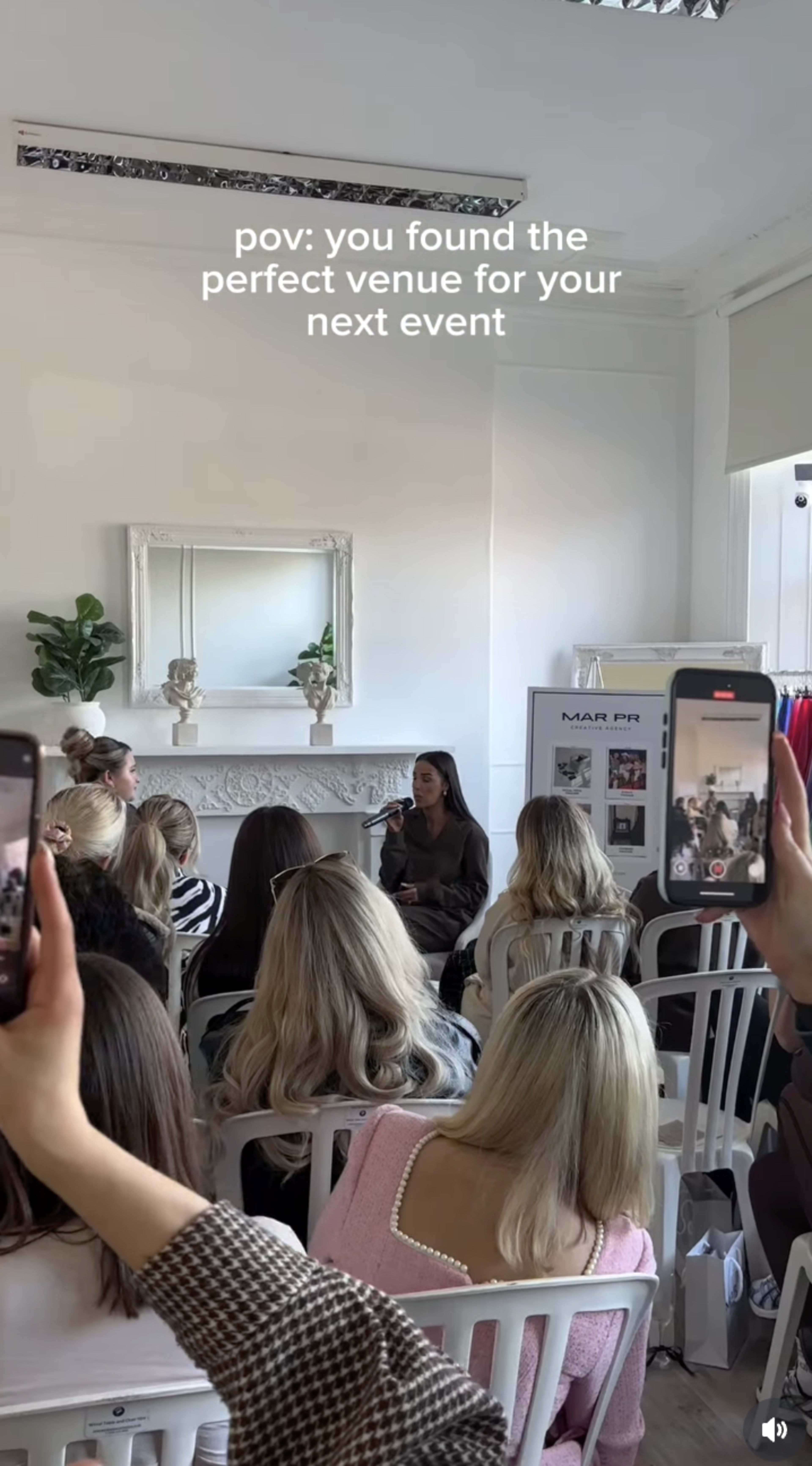 A group of attendees sits in a well-lit room, facing a speaker while others hold up their phones to capture the moment.