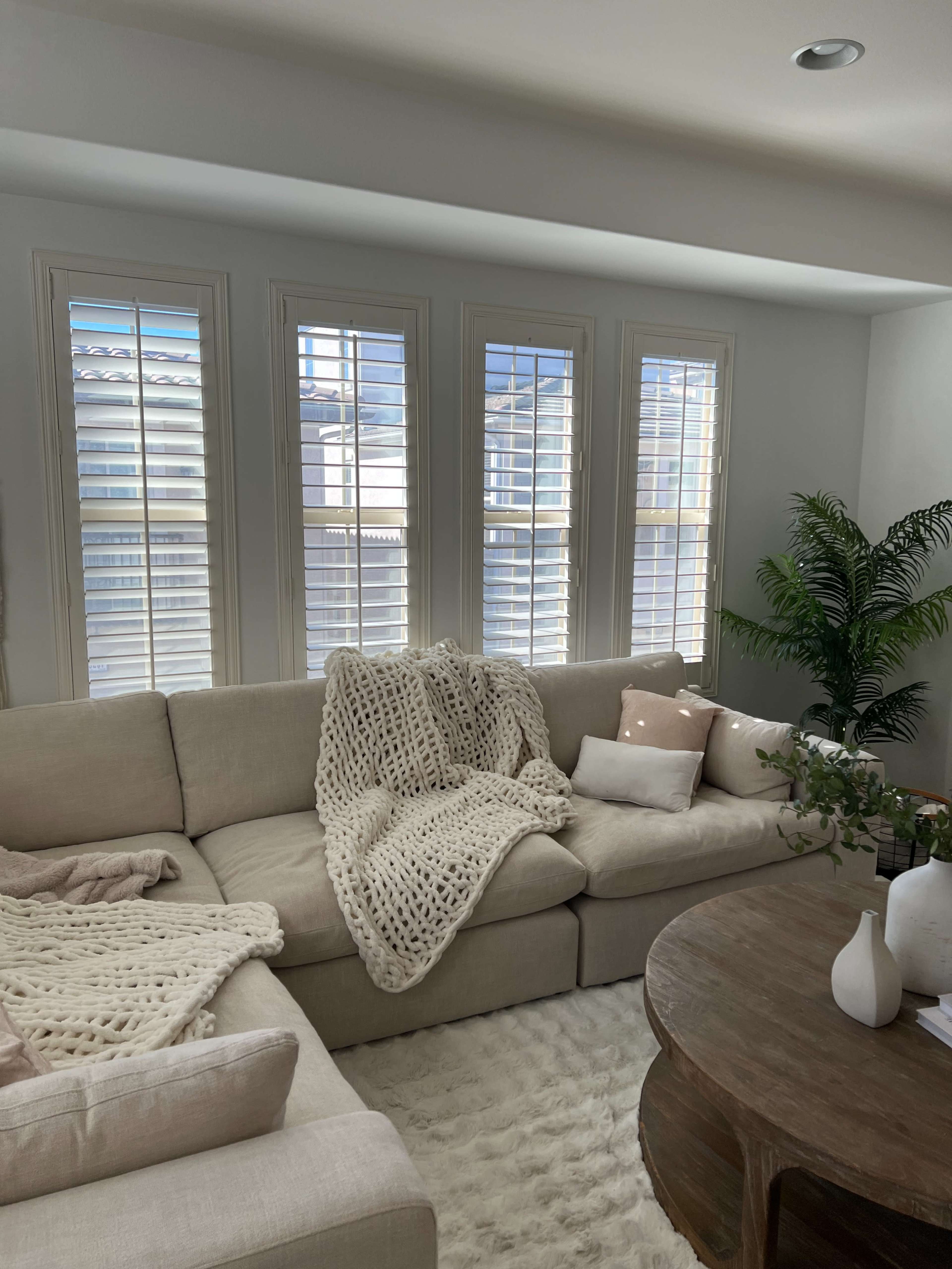 A light-colored sectional sofa is positioned in a cozy living room, adorned with a chunky knit throw, a round wooden coffee table, and large windows with plantation shutters.