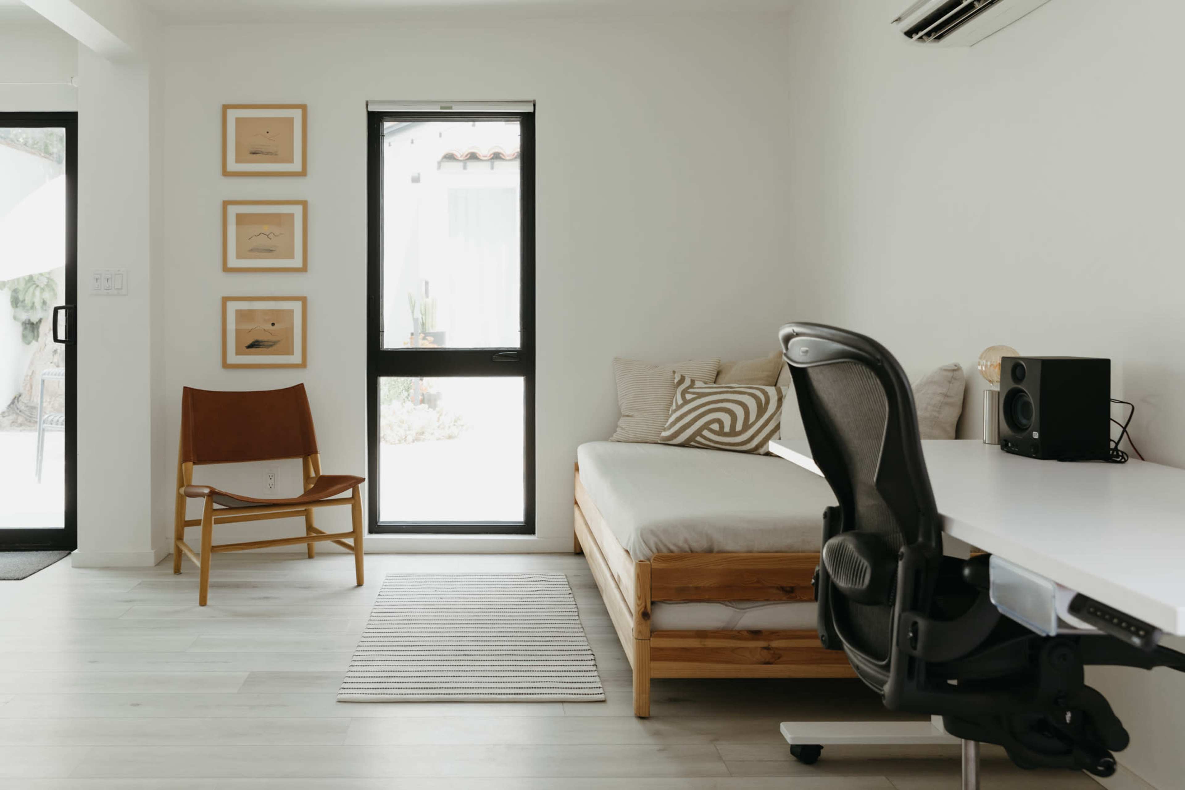 A minimalist living space with a wooden daybed, a wooden chair, and a desk with a speaker, all situated near large windows that let in natural light.