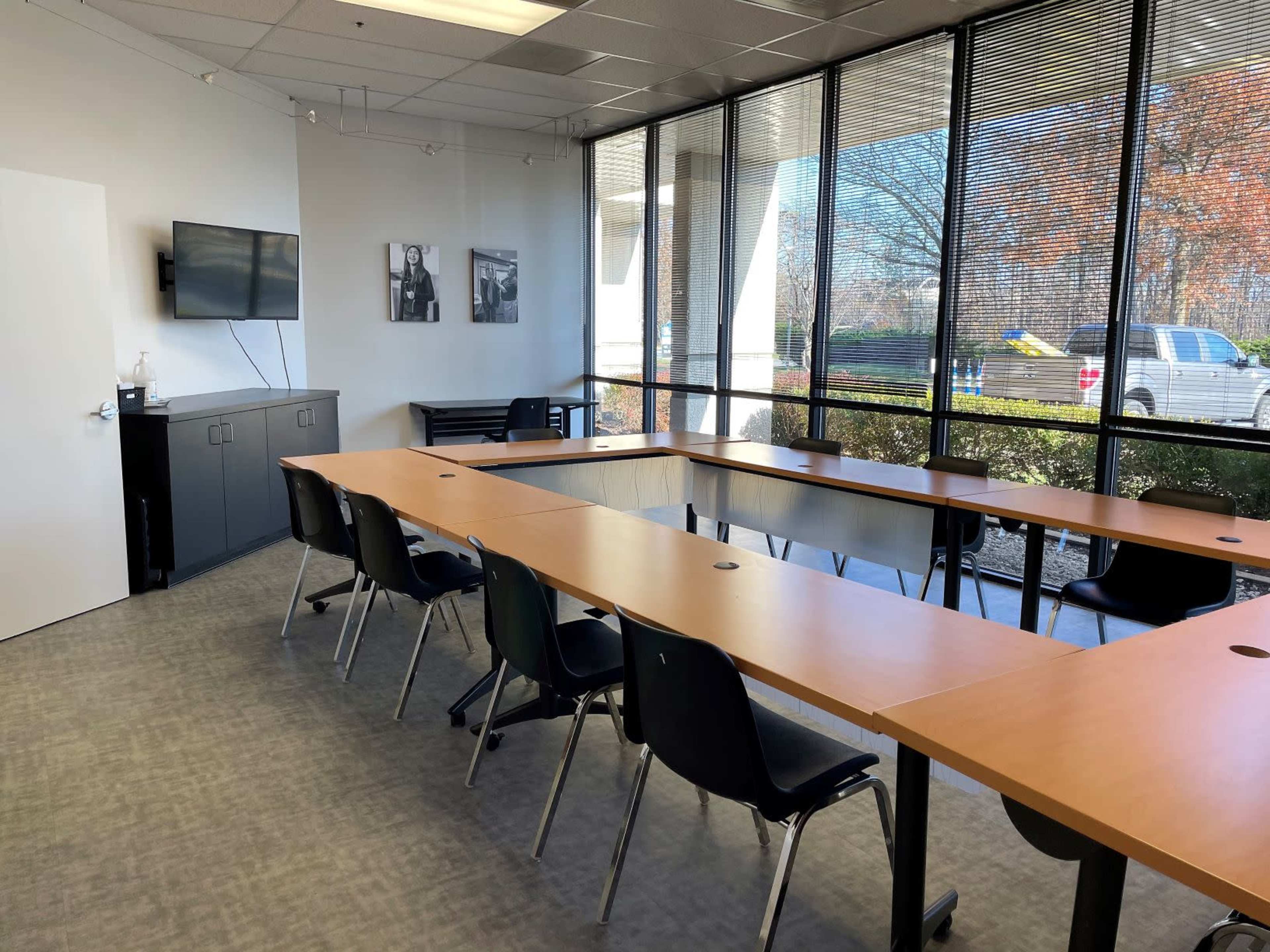 A bright conference room features a long wooden table with black chairs, large windows providing natural light, and a television mounted on the wall.