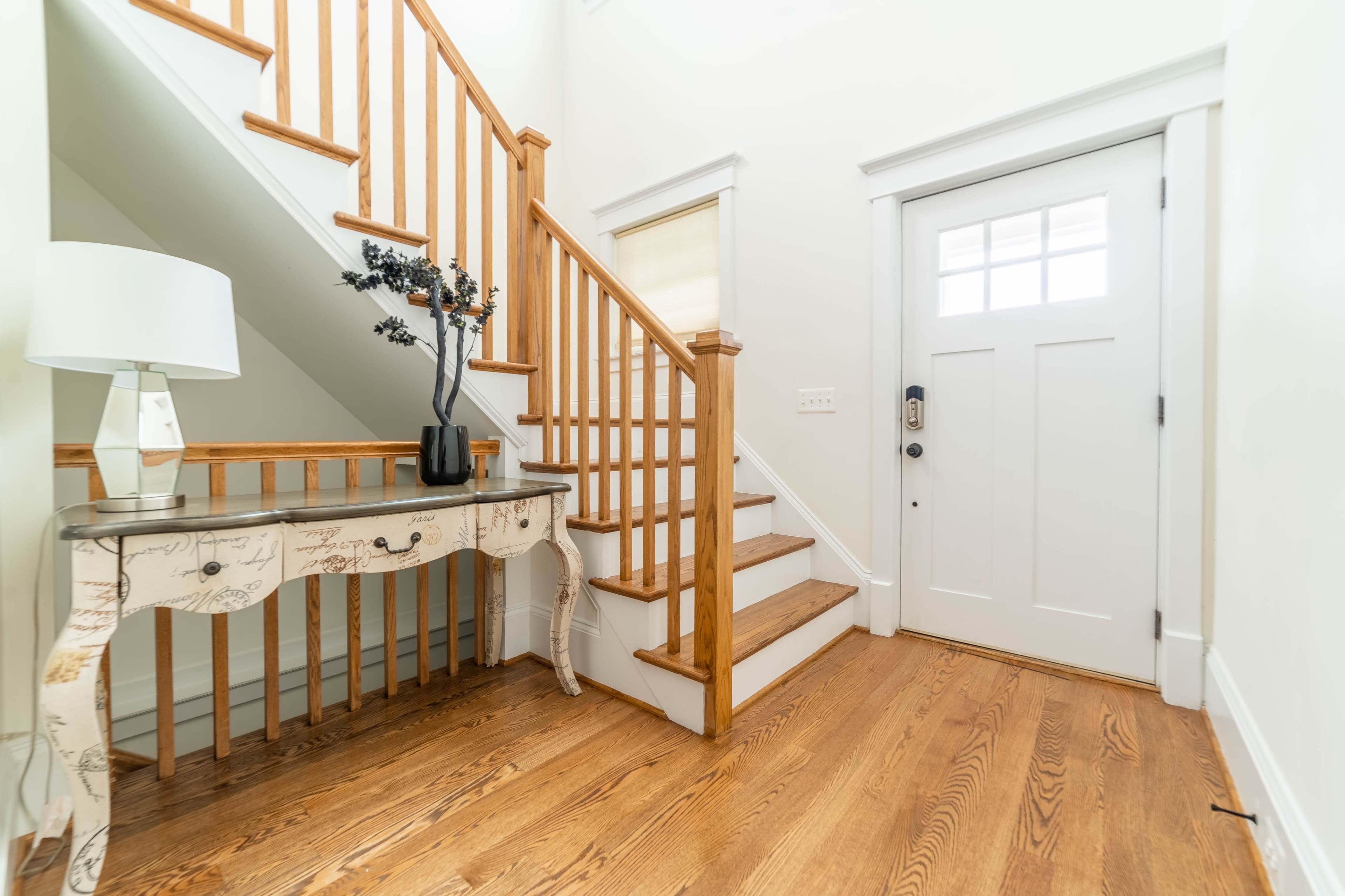 A well-lit entryway features a wooden staircase, a console table with a vase, and a front door with multiple glass panels.