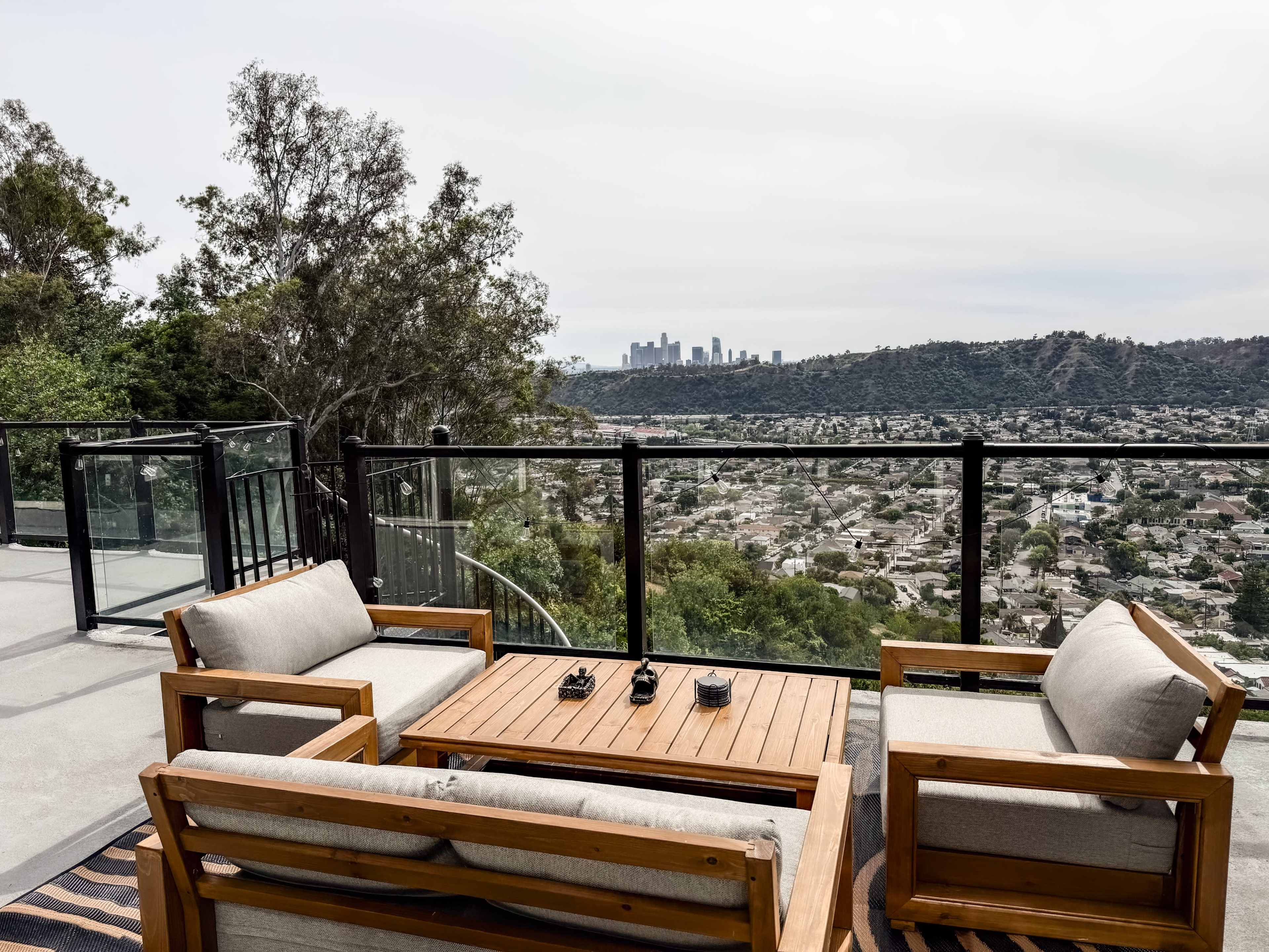 The image shows a modern outdoor seating area with wooden furniture overlooking a city skyline from elevated terrain.