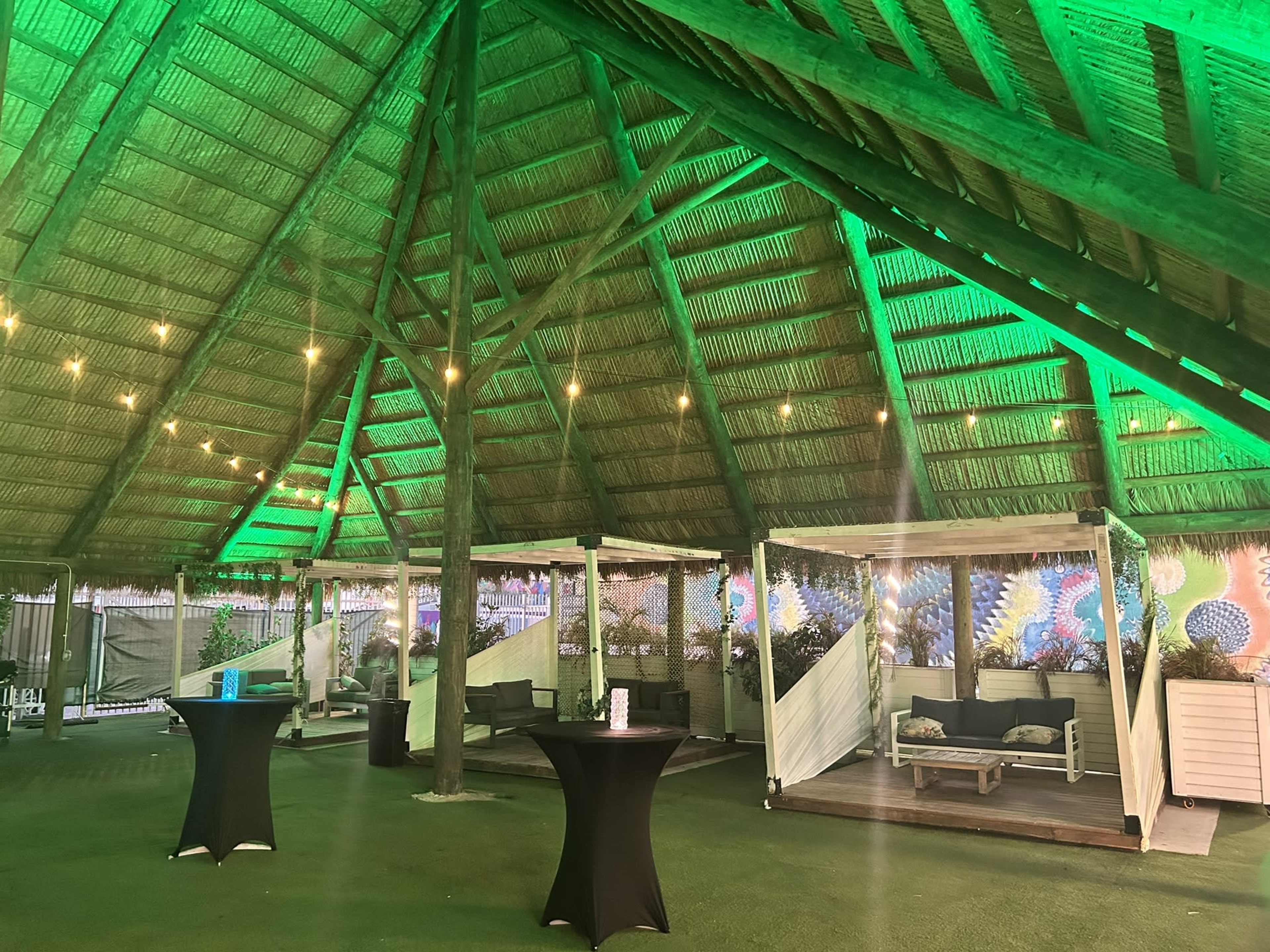 The image shows a spacious indoor area with a thatched roof, featuring green lighting, several small tables, and lounge seating.