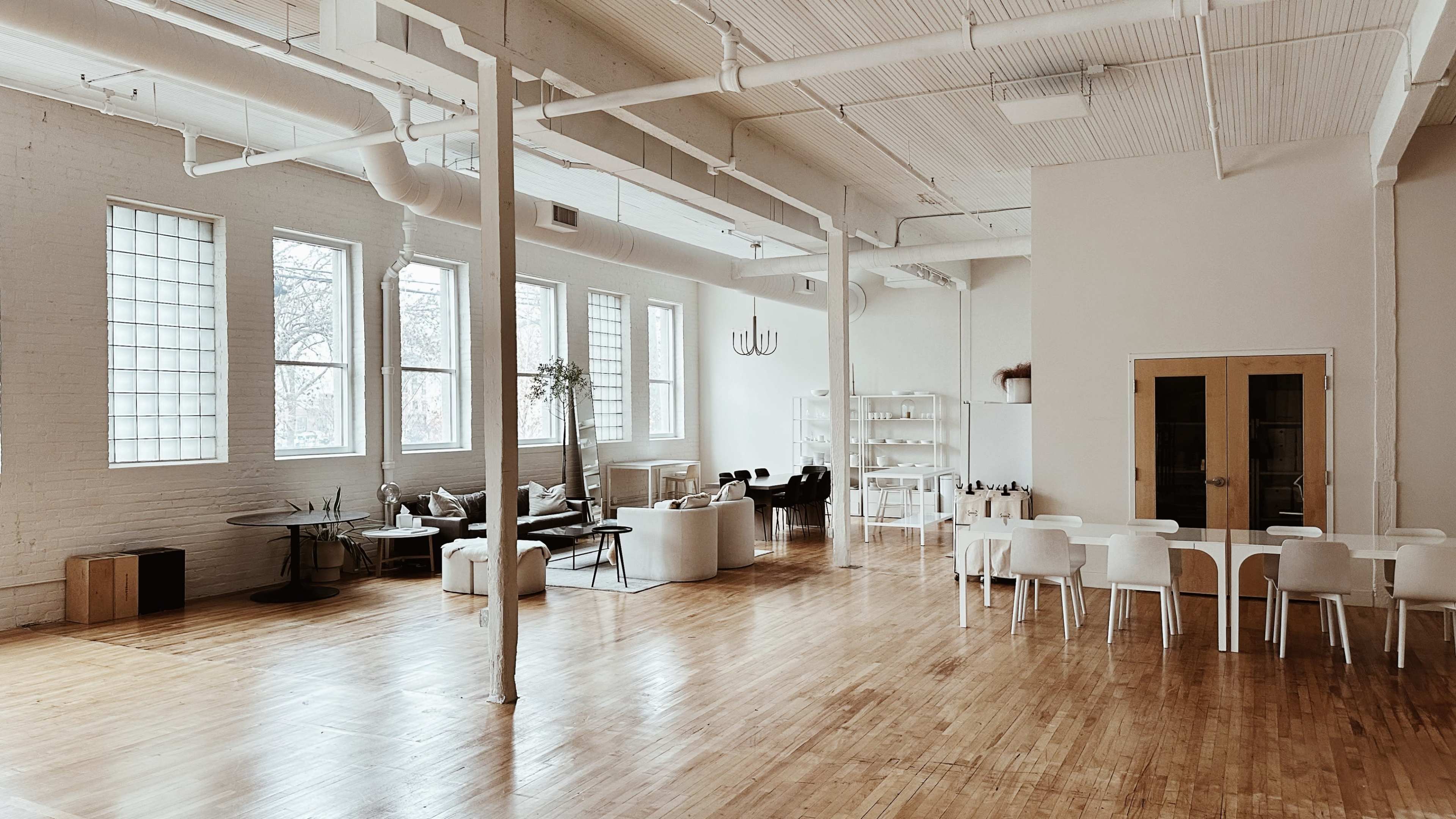 A spacious, well-lit interior of a modern, minimalist room featuring white walls, large windows, various seating areas, and wooden flooring.