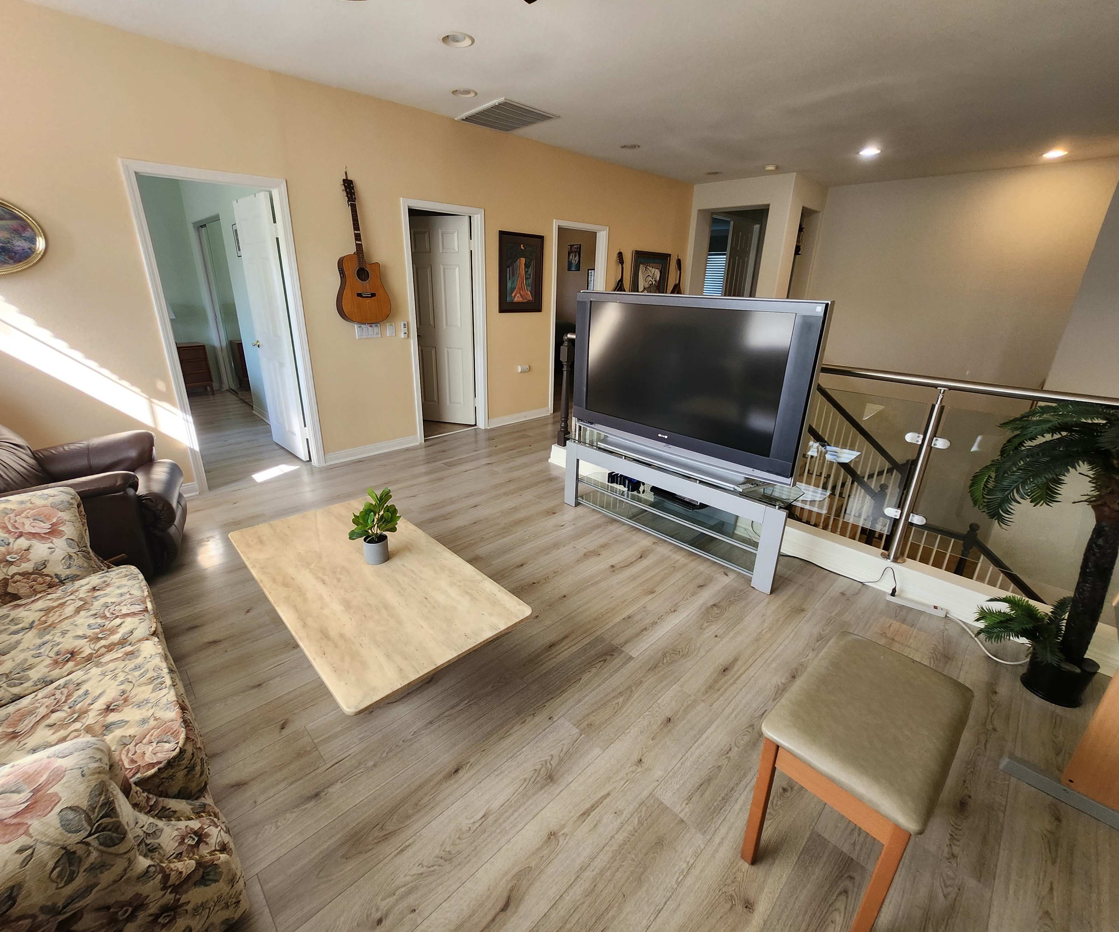 The living room features a floral-patterned sofa, a large TV on a stand, a coffee table, and a chair, with doors leading to other rooms.