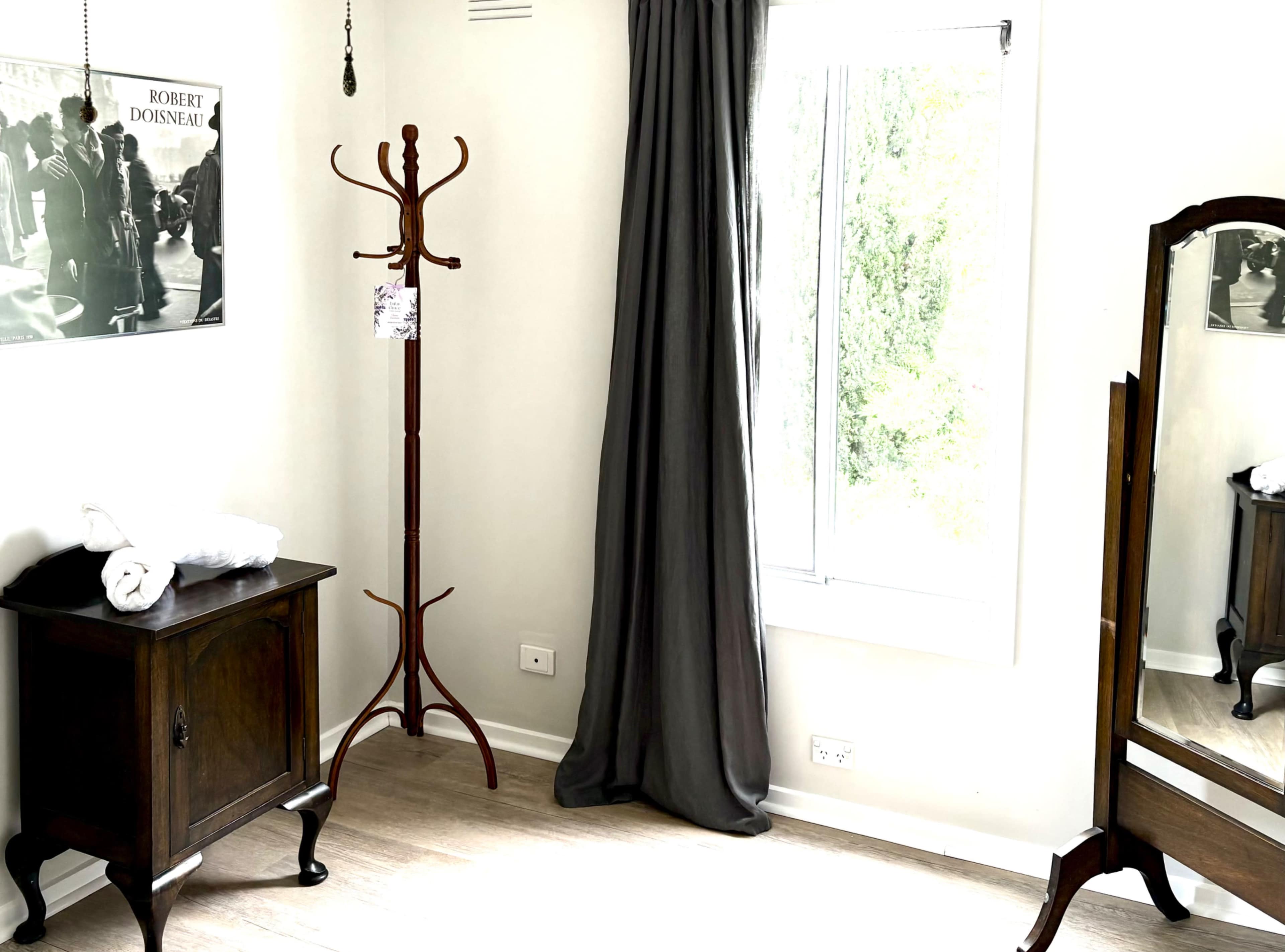 The image shows a minimalist bedroom corner featuring a wooden coat rack, a vintage dresser, a full-length mirror, and a window with grey curtains.