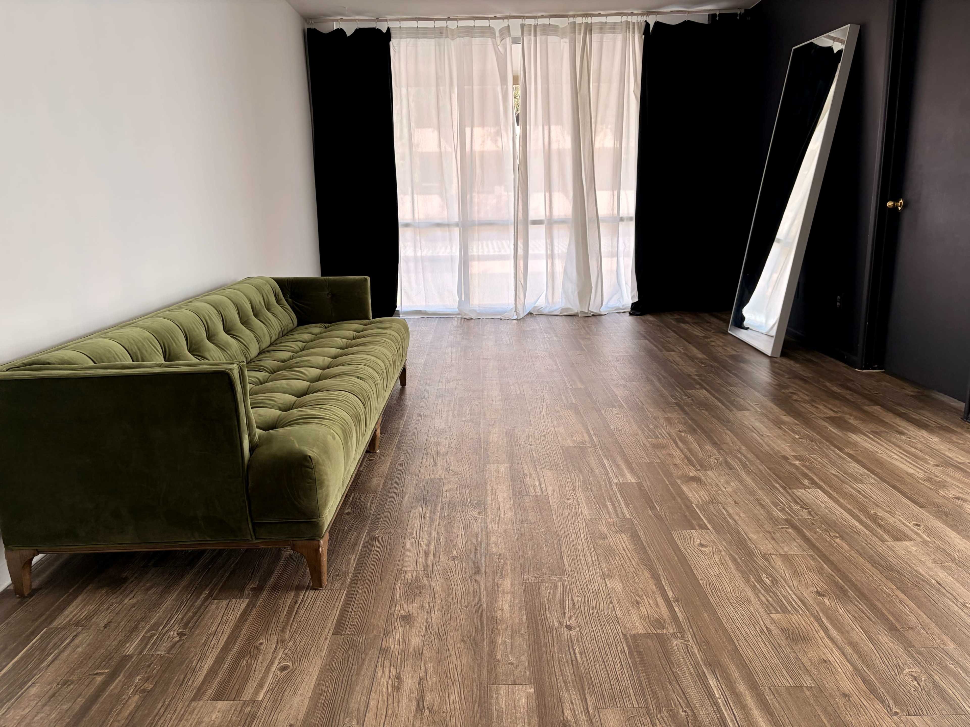 The image shows a spacious room with a green couch, large windows covered by sheer curtains, and a full-length mirror against a black wall.