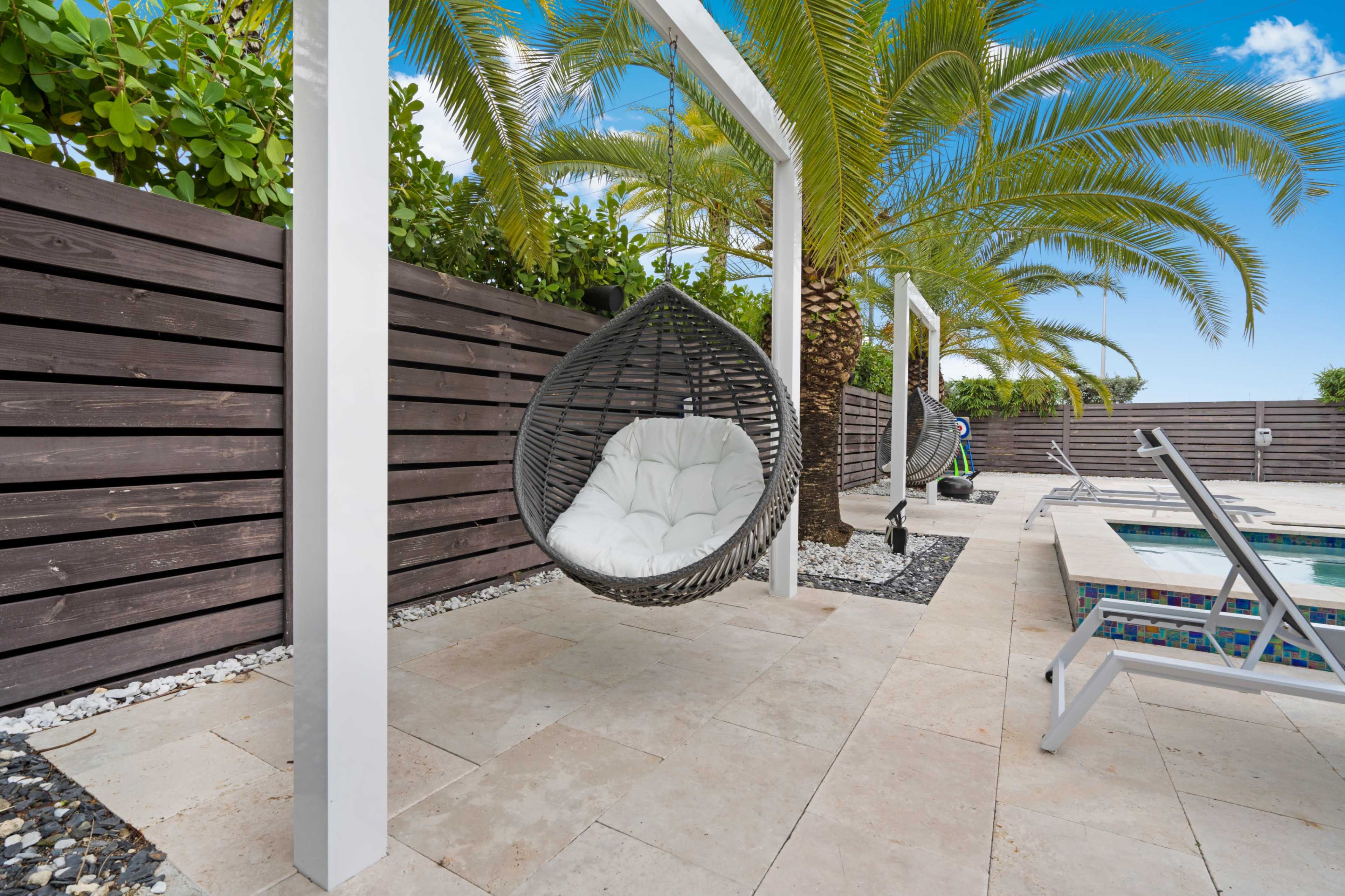 A hanging chair is suspended from a white frame beside a swimming pool, surrounded by palm trees and a wooden fence.