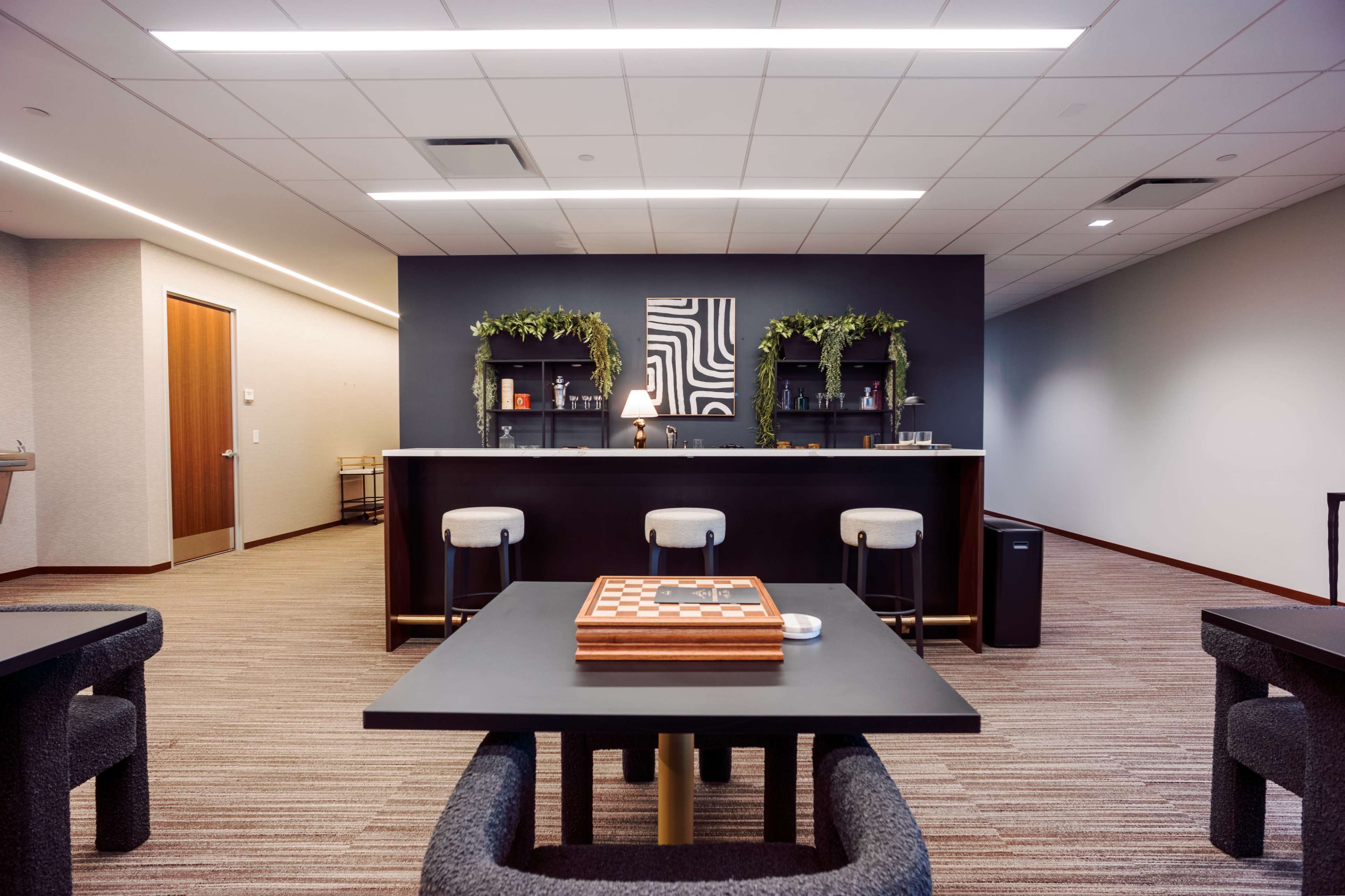 The image shows a spacious lounge area featuring a bar with shelves and a few tables, decorated with greenery.