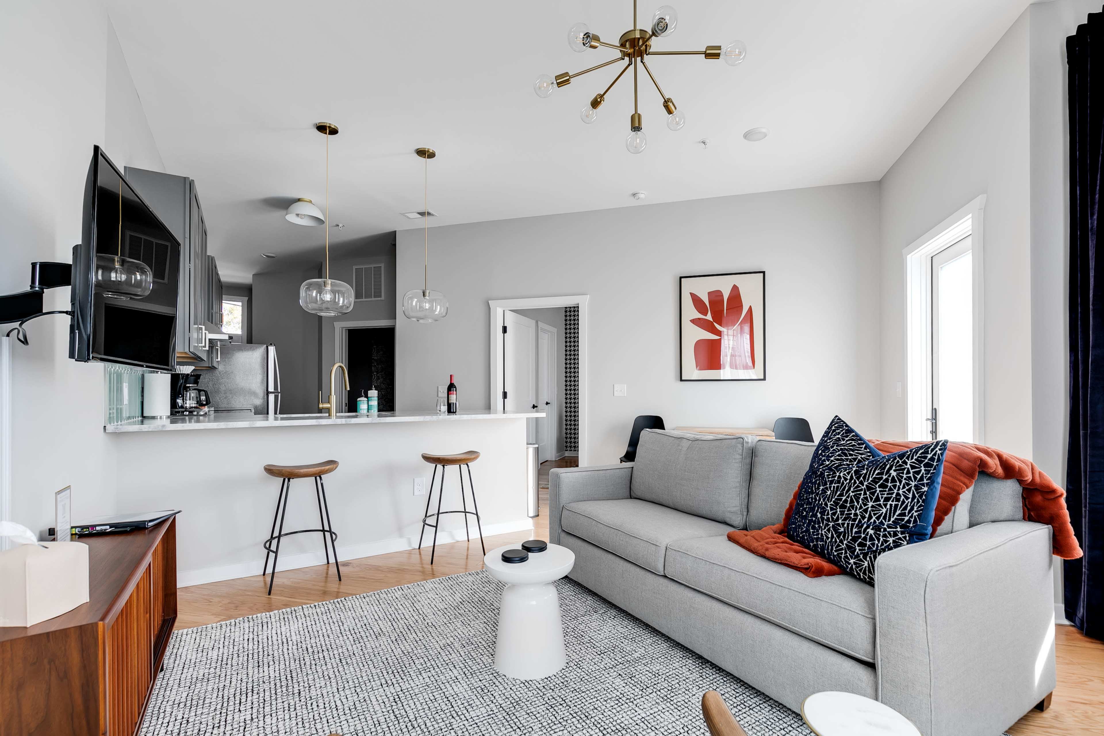 A modern living room featuring a gray sofa, a round coffee table, a wall-mounted television, and a kitchen area with a bar counter.